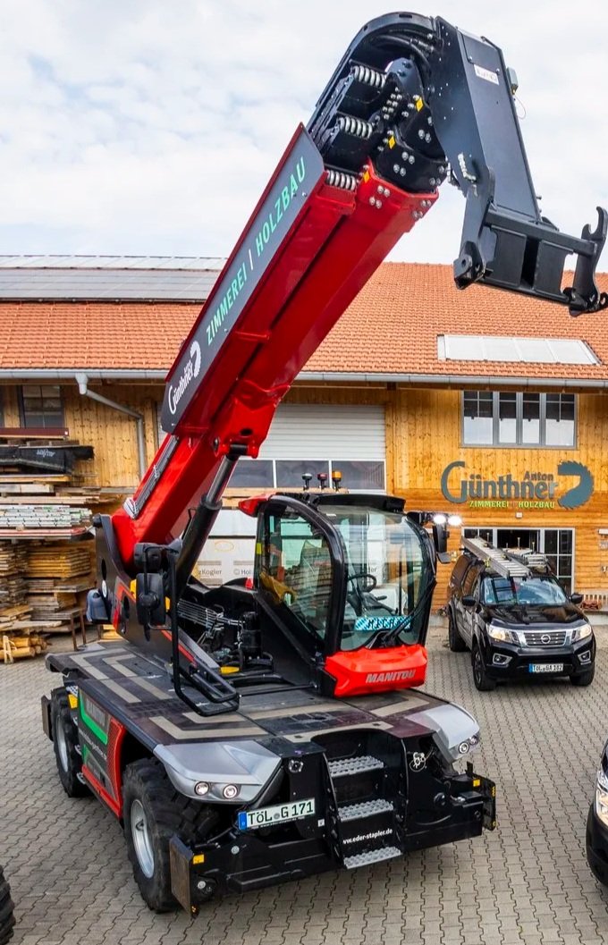 Ein roter Teleskopkran auf einem gepflasterten Parkplatz vor einem Holzhaus mit Schindeldach. Es gibt zwei Autos und ein Gebäude mit Schaufenster und einer Beschilderung auf dem Hintergrund.