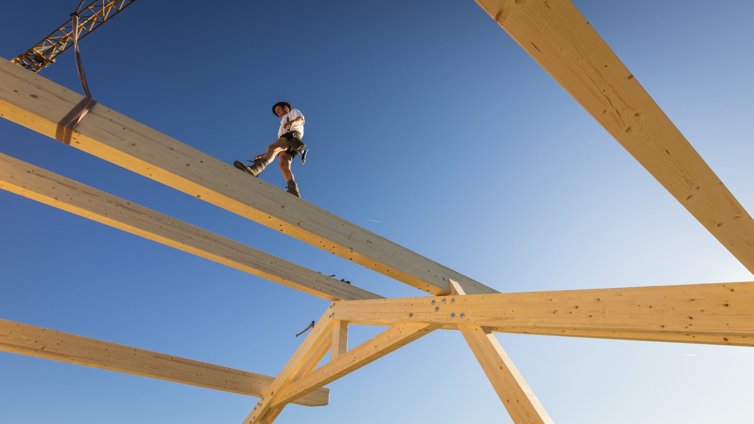 Ein Mann arbeitet an einem Holzrahmen unter klarem blauen Himmel, während er auf den Holzbalken steht.