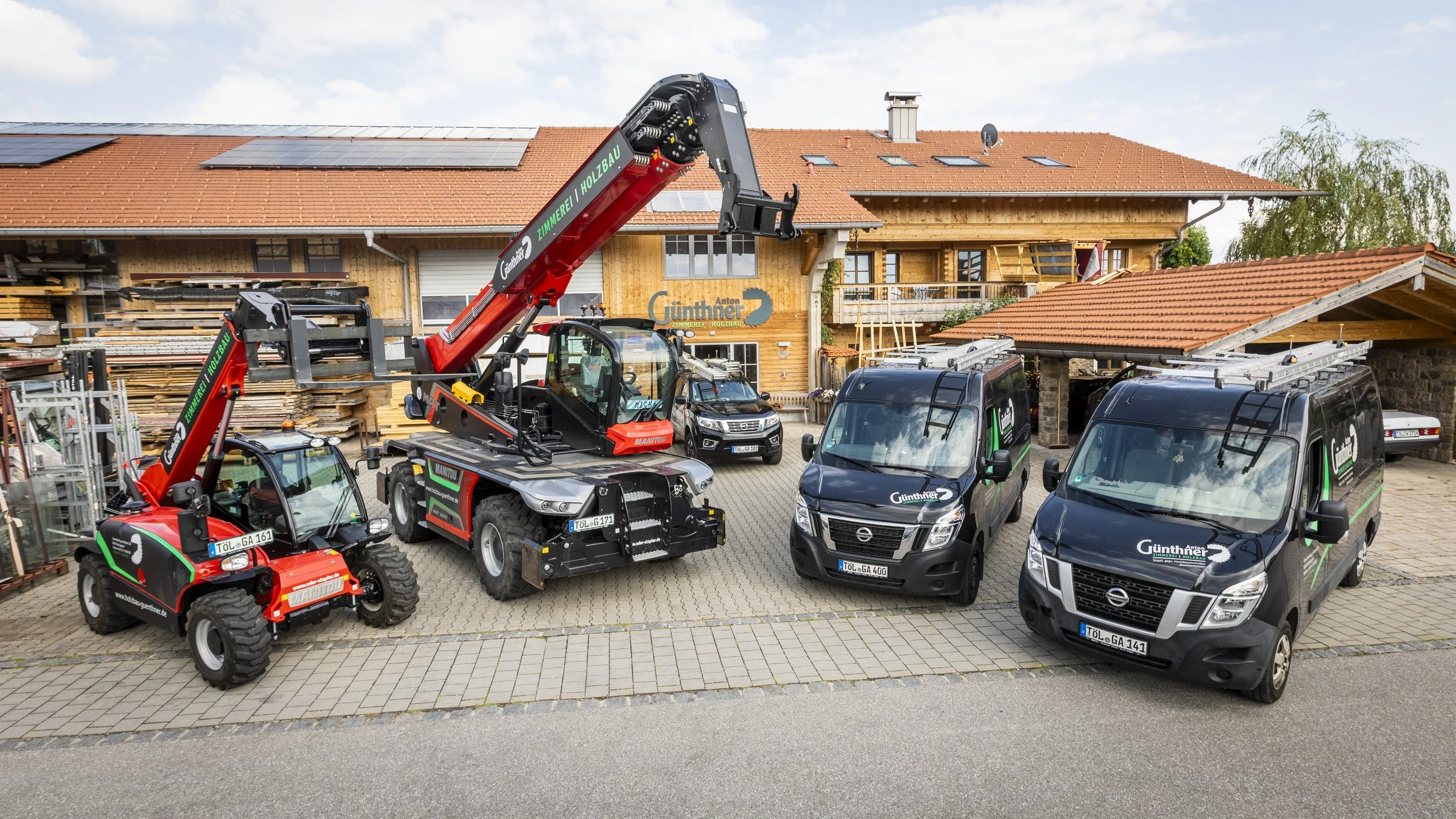 Bauernhof mit Baumaterialien, Baumaschinen, und Fahrzeugen vor einem Holzhaus mit roten Dachziegeln und Solarpanelen.