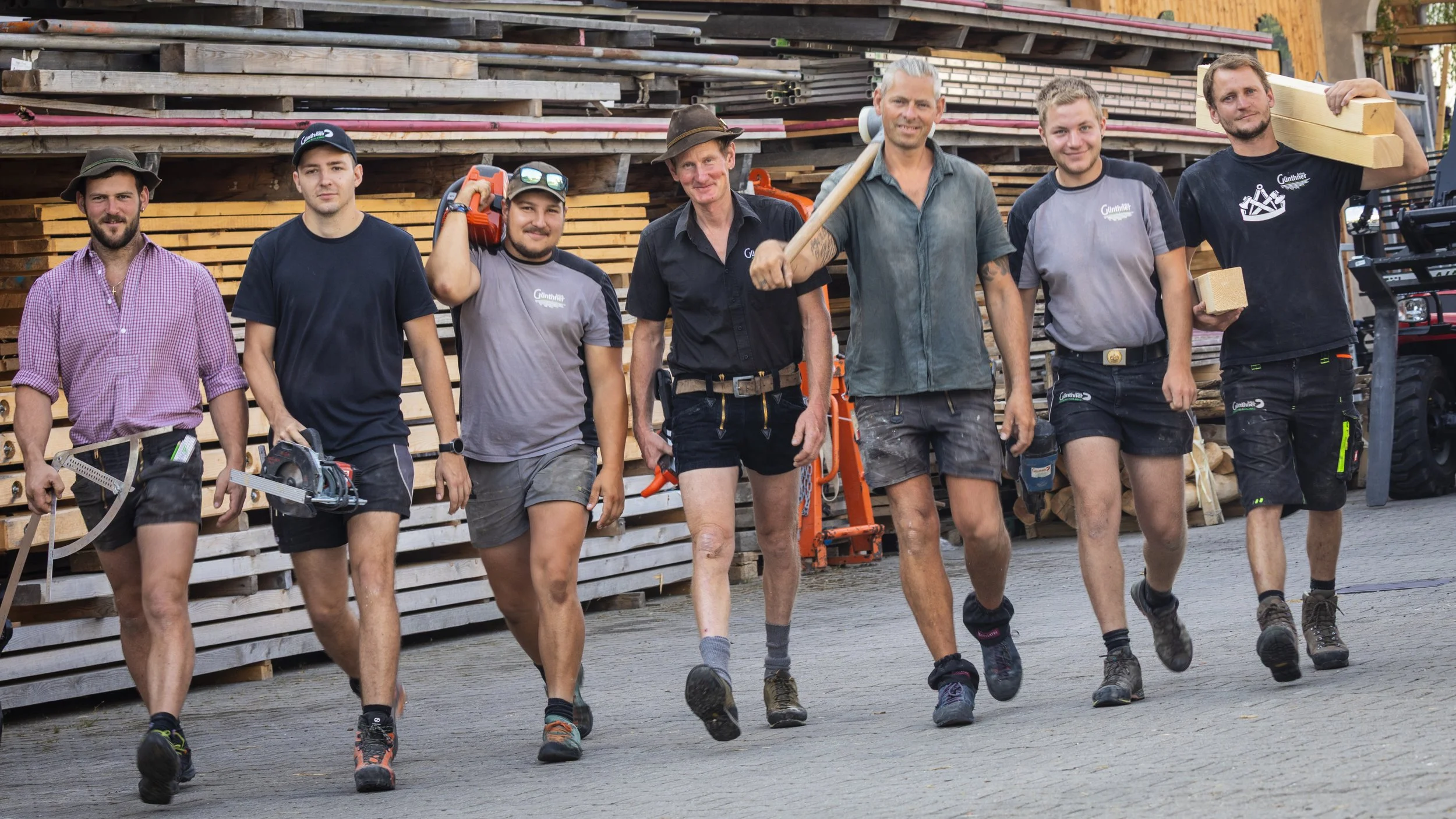 Sieben Männer in Arbeitskleidung laufen zusammen vor einem Stapel Holz, einige tragen Werkzeuge und Ausrüstung. Es sieht nach einem Bau- oder Handwerksbetrieb aus.