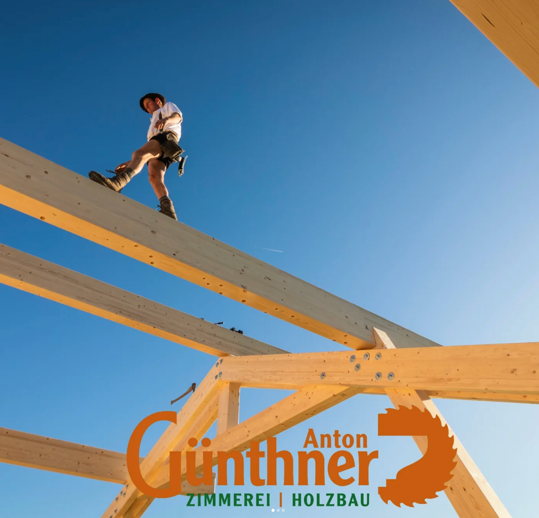 Blick auf den Rahmen eines Holzhauses unter blauer Himmel, mit sichtbaren Holzbohlen und Holzbalken. Der Rahmen ist Teil eines Bauprojekts im Rohbau. Text im unteren Bereich in orange und grün, mit Firmenname und Tätigkeitsbeschreibung.