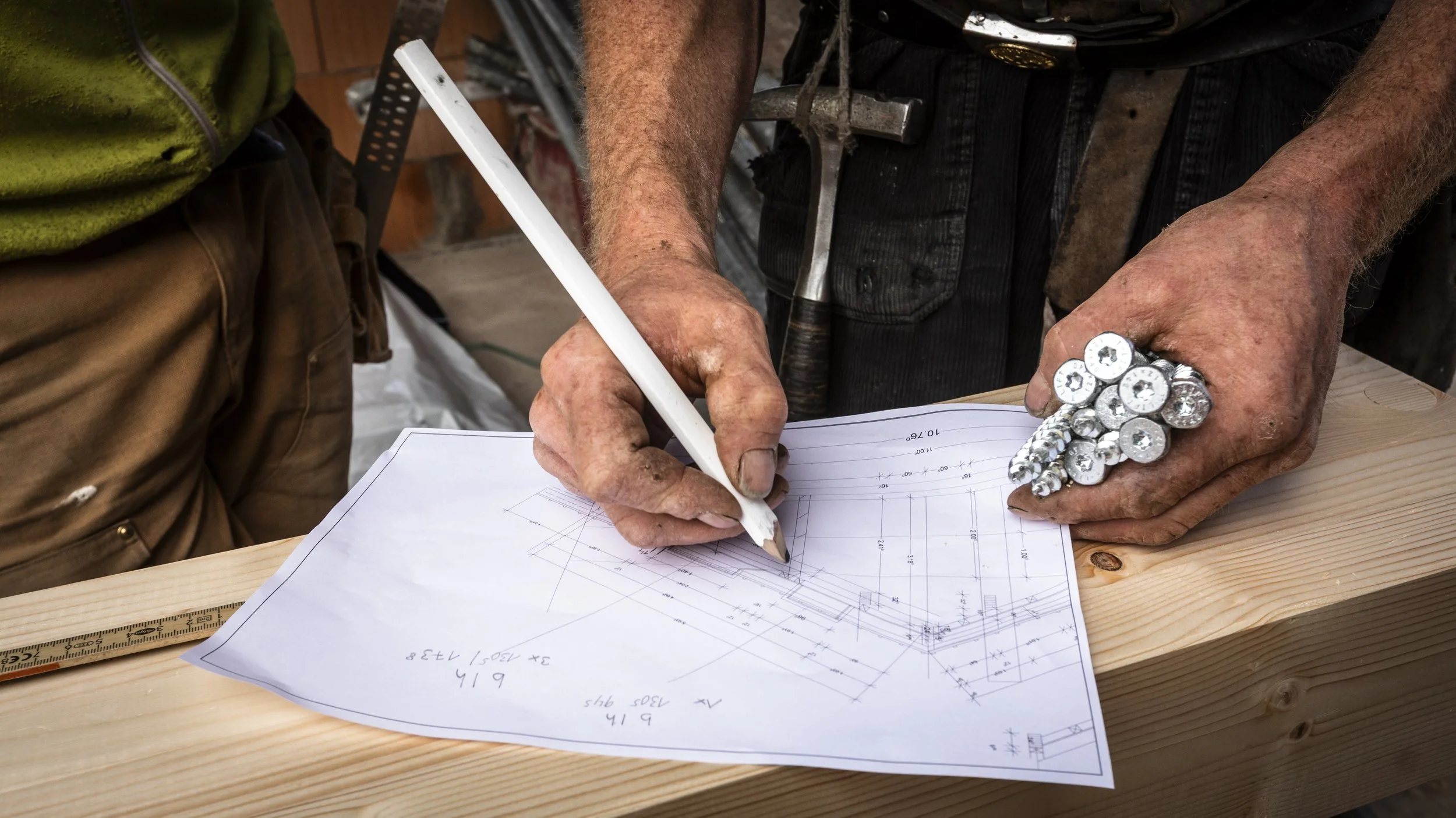 Ein Handwerker, der auf einen technischen Bauplan auf einem Holzbrett zeichnet, trägt einen großen Ring aus Schrauben am Finger und hat ein Werkzeug in der Tasche.