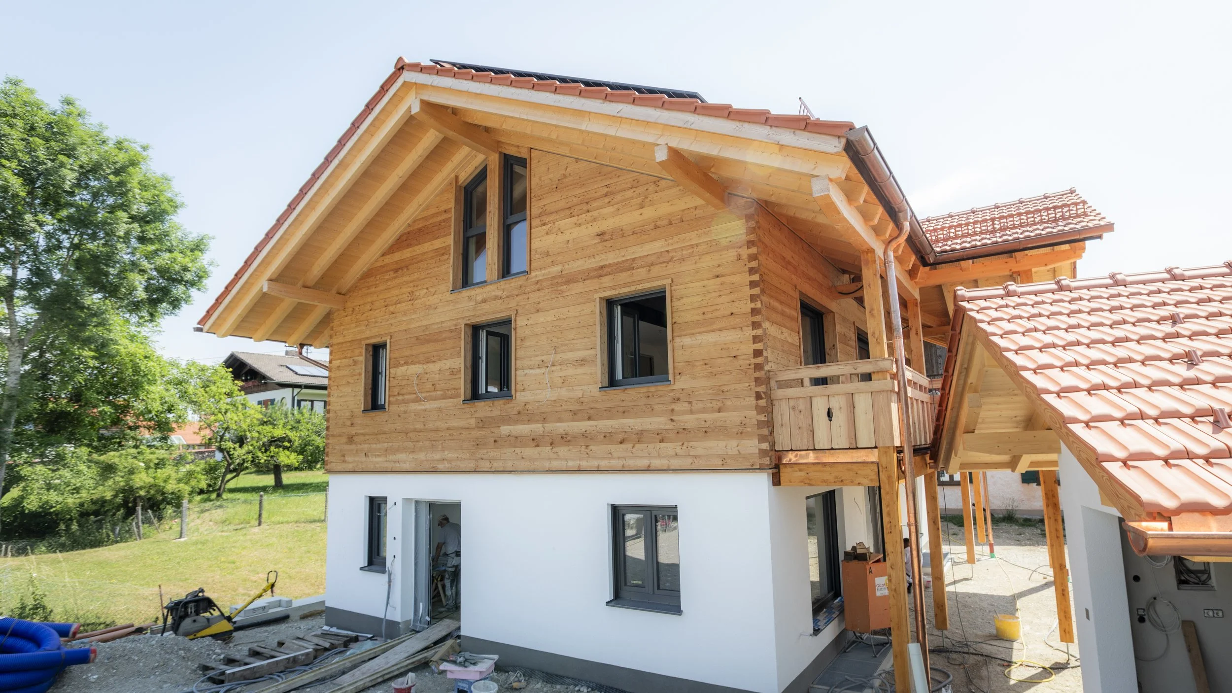 Ein Holzhaus im Bau mit offener Fensterfronten und roten Dachziegeln, umgeben von Grünflächen und einer Baustelle im Vordergrund.
