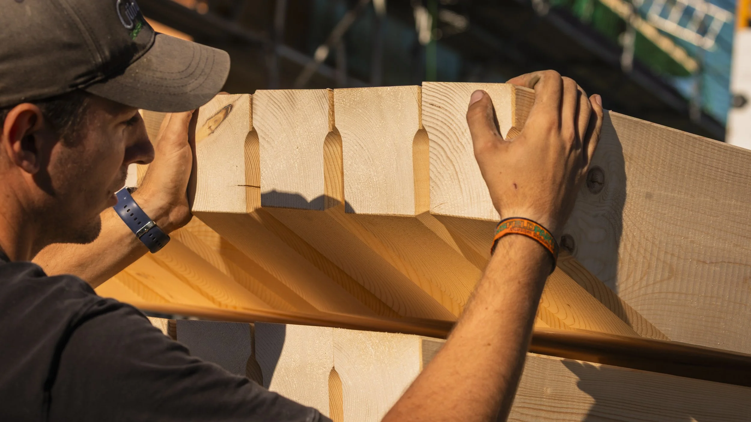 Nahaufnahme eines Mannes, der an einem Holzbalken arbeitet. Er trägt eine schwarze Mütze, einen schwarzen T-Shirt und einen blauen Armband. Die Hände sind schmutzig, während er die Holzstücke anfasst, passend für eine Bau- oder Holzbearbeitungsarbeit.