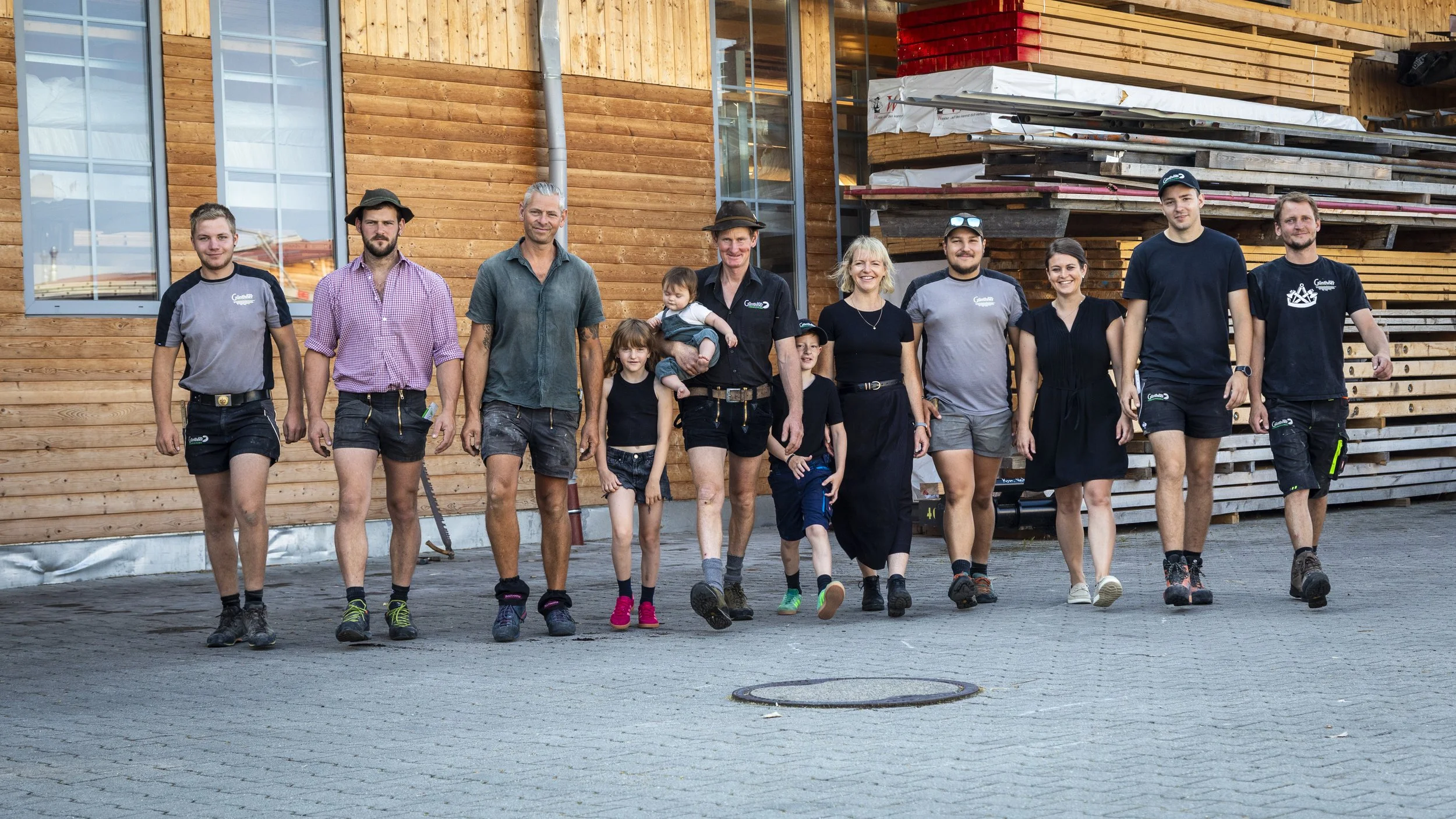 Gruppe von Menschen, darunter Kinder und Erwachsene, beim Spaziergang vor einem Holzhaus, im Hintergrund Baustoffe und Baugerüst