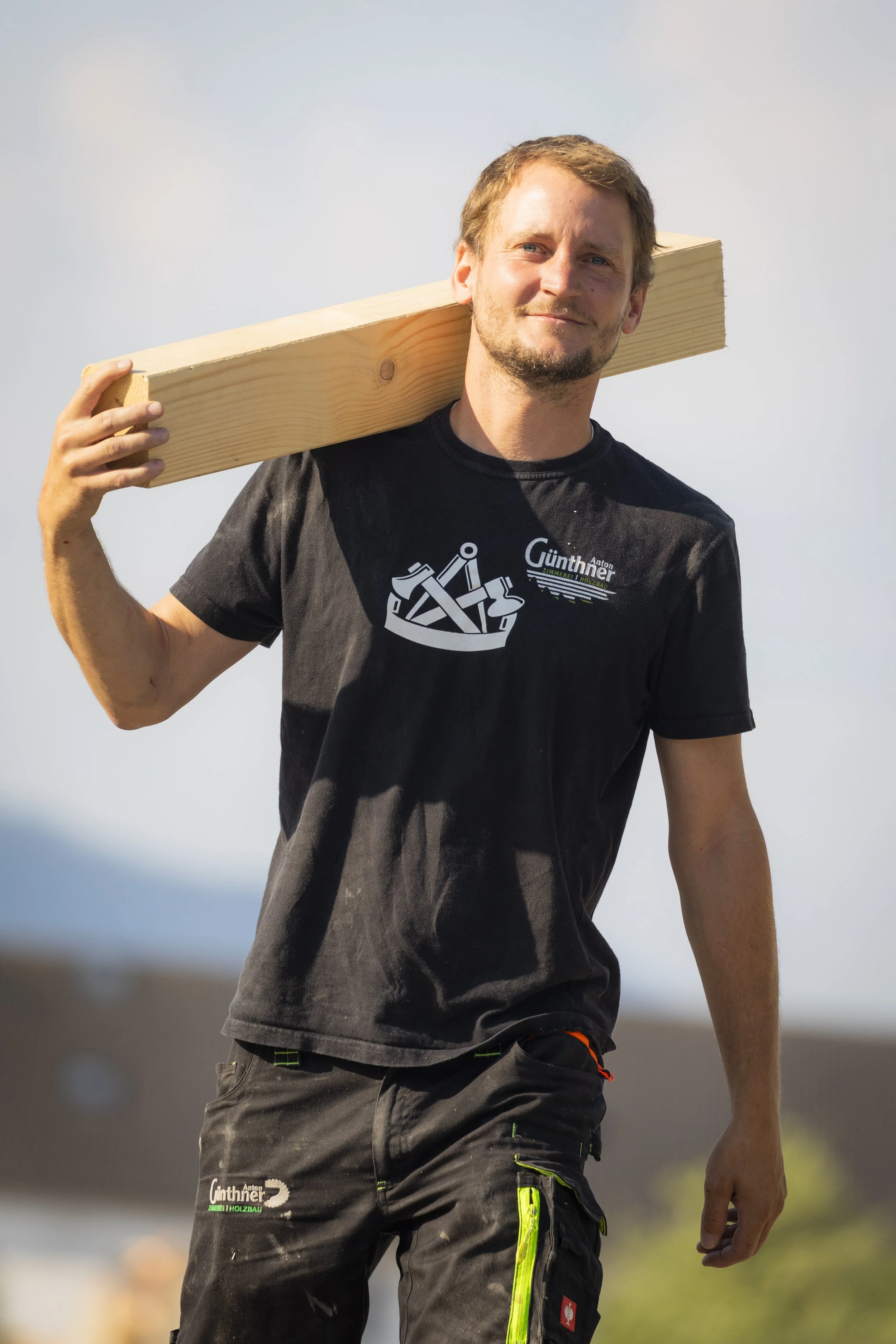Junger Mann trägt schwarze Arbeitshose mit Neon-Details und schwarzen T-Shirt, hält ein Holzbalken auf Schulter, Outdoor-Hintergrund, leichter Himmel.