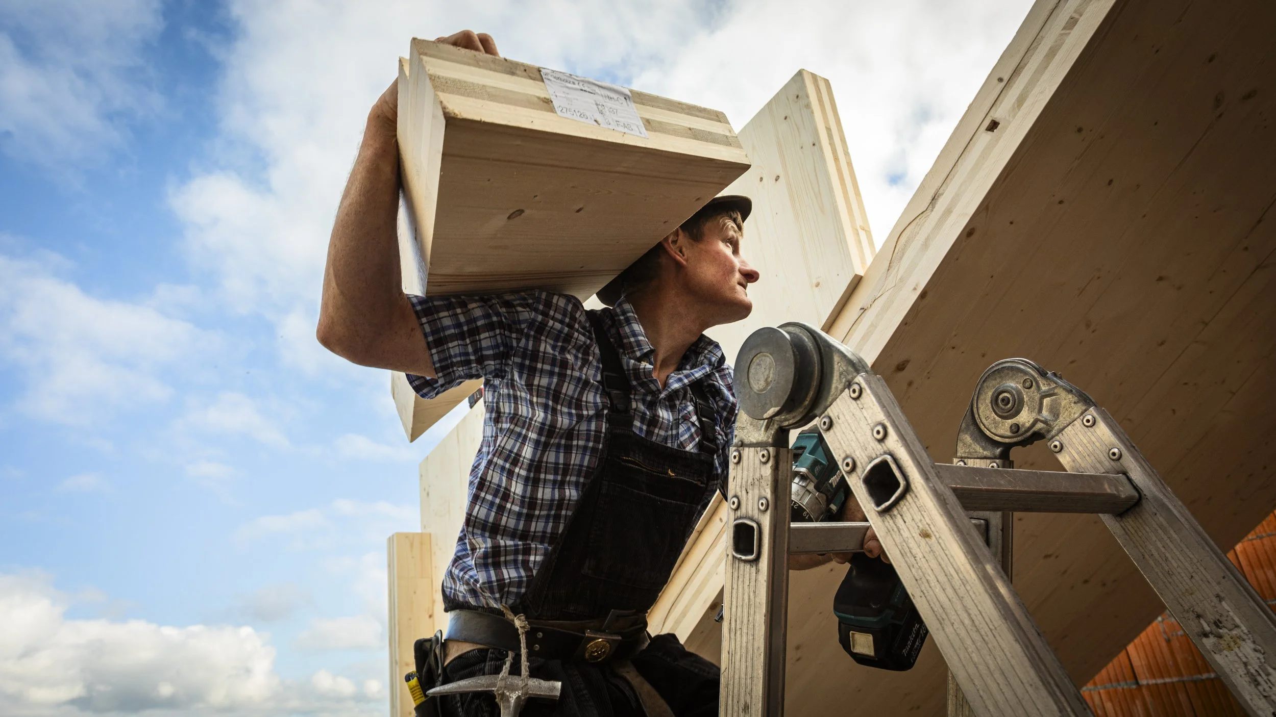 Handwerker auf einer Leiter beim Holzbau, trägt Holzbrett auf der Schulter, blauer Himmel mit Wolken im Hintergrund.
