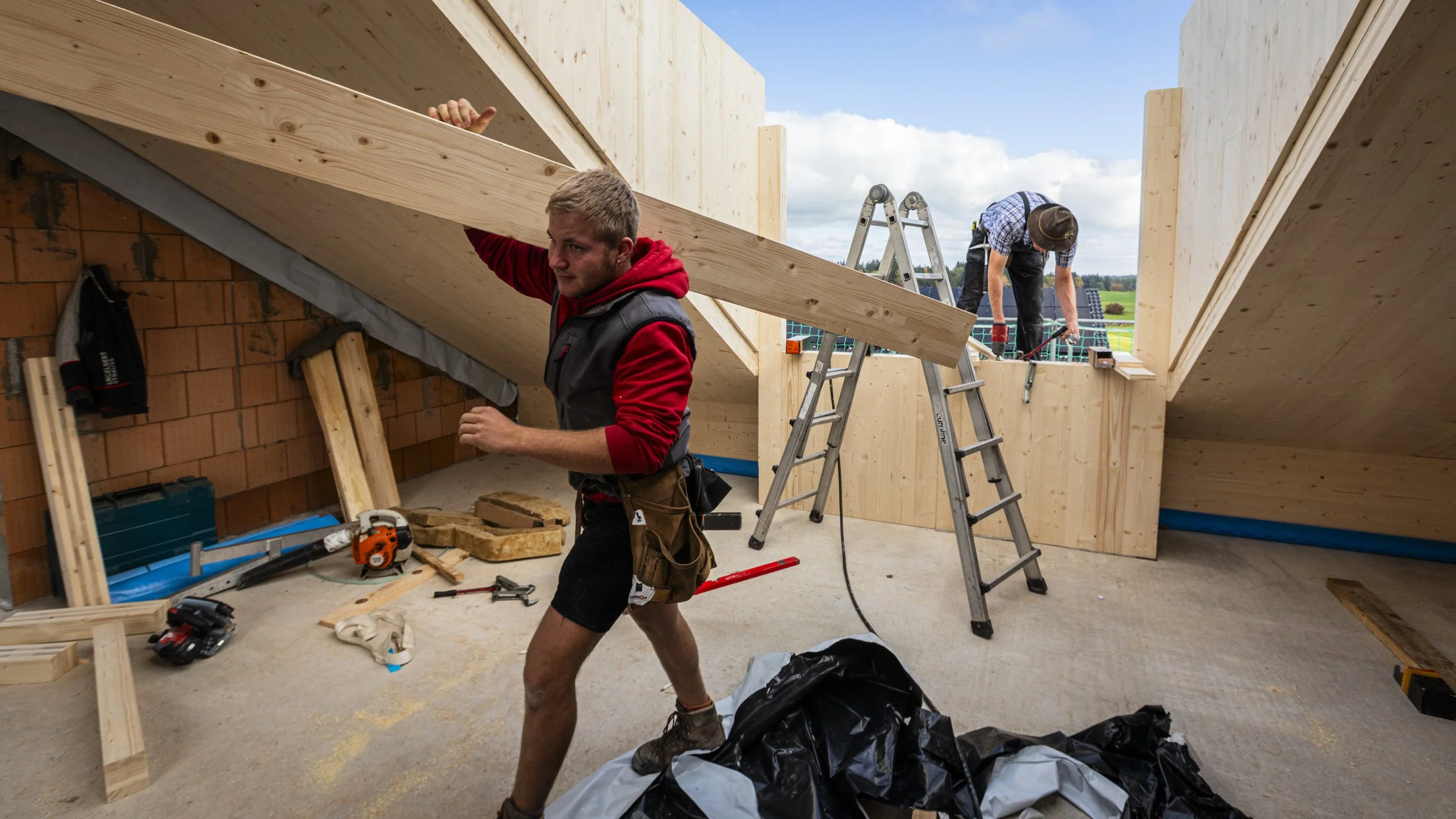 Zwei Bauarbeiter installieren eine Dachkonstruktion aus Holz in einem noch im Bau befindlichen Haus, in der Umgebung sind grüne Felder und Himmel zu sehen.