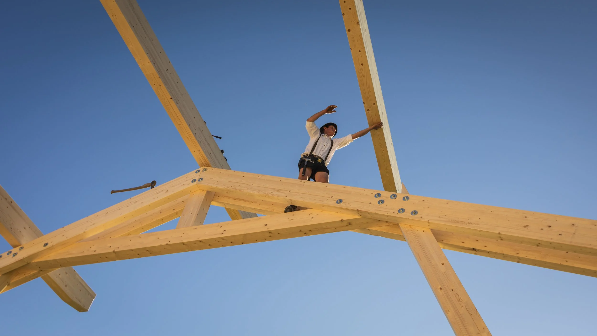 Ein Mann steht auf einem Holzbau, vermutlich beim Bau eines Hauses, und streckt die Arme in die Luft, während er in den Himmel blickt. Das Bild ist gegen den blauen Himmel aufgenommen.