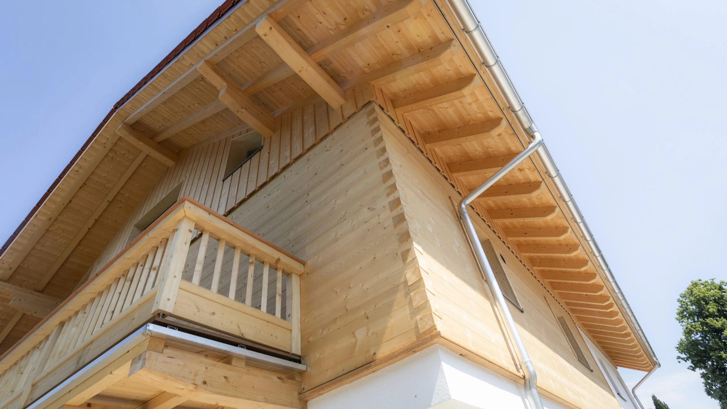 Holzhaus mit Balkon und Dachüberstand, Blick nach oben, blauer Himmel, eine Baumkrone ist sichtbar.