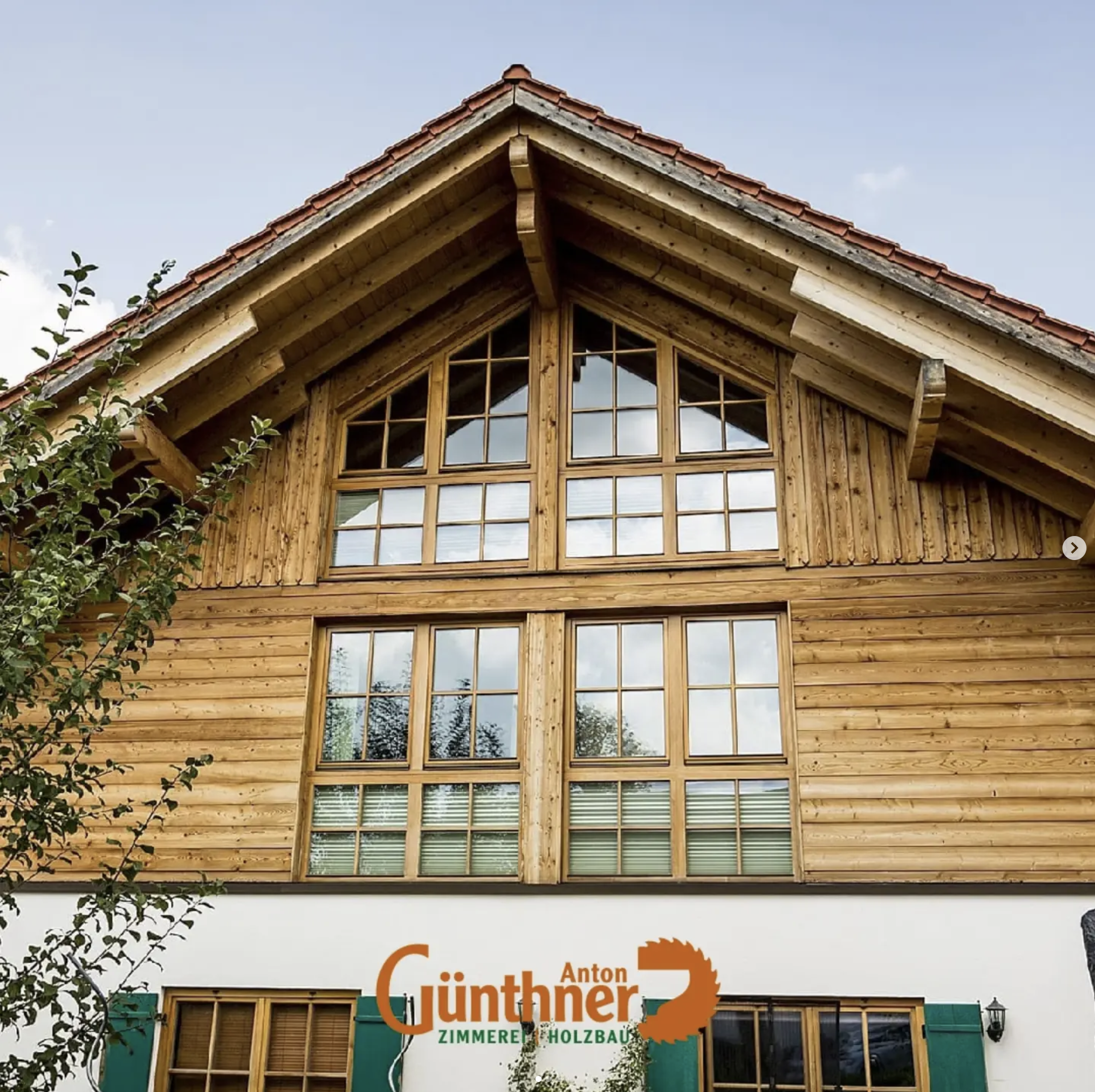 Das Bild zeigt das Dach eines Holzhauses mit großen Fenstern und einem Firma-Logo auf der unteren Fassade. Der Himmel ist leicht bewölkt.