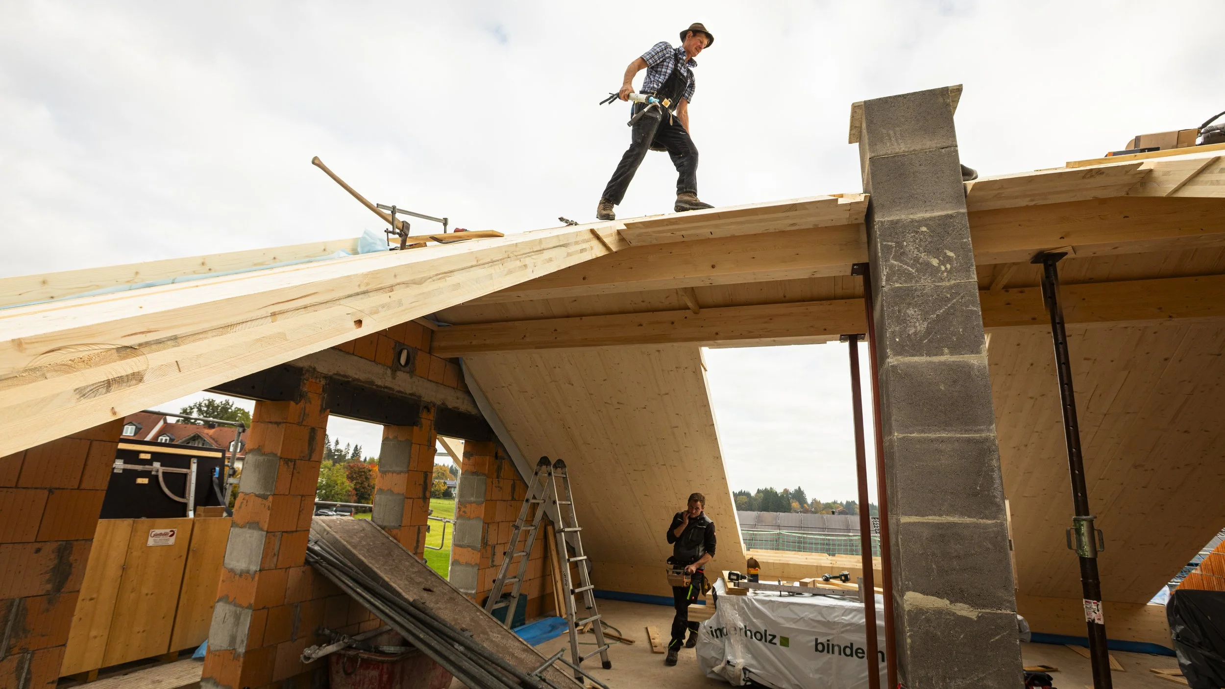 Ein Bauarbeiter steht auf einem Holzbaugerüst und arbeitet an einem Dach, während ein Kollege im Innenraum das Bauprojekt überwacht.