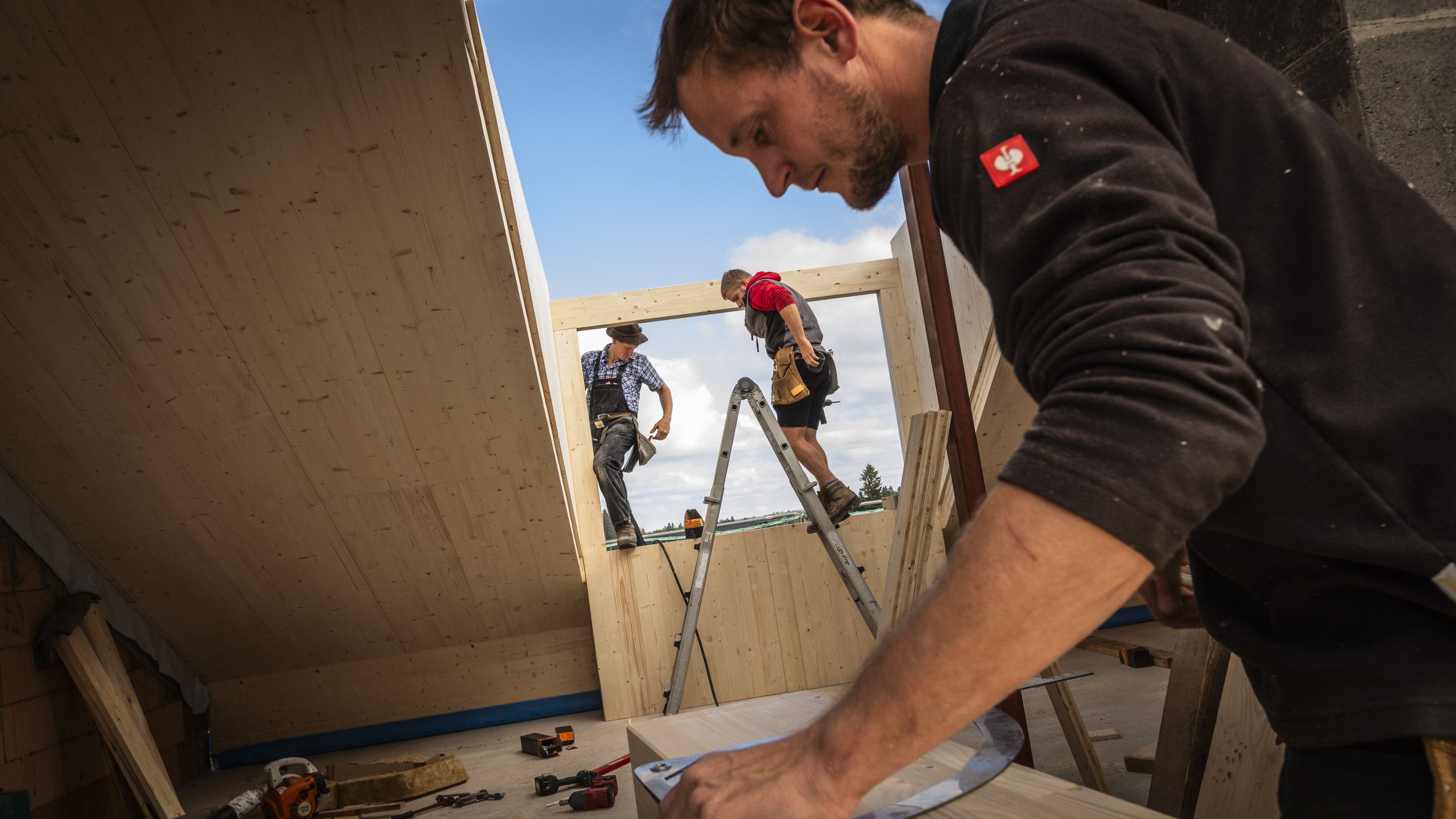 Bauarbeiter arbeiten an einem Holzrahmen eines Gebäudes, während ein Mann im Vordergrund eine Werkzeuge benutzt, mit Werkzeugen auf dem Boden neben sichche