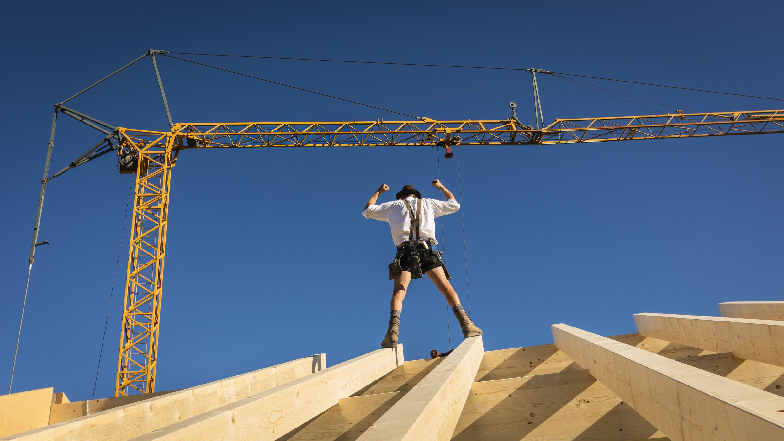 Ein Bauarbeiter steht auf einem Holzrahmen auf einer Baustelle und trägt Sicherheitsgurte, während er die Holzbalken befestigt. Im Hintergrund ist ein Kran sichtbar, und der Himmel ist klar und blau.