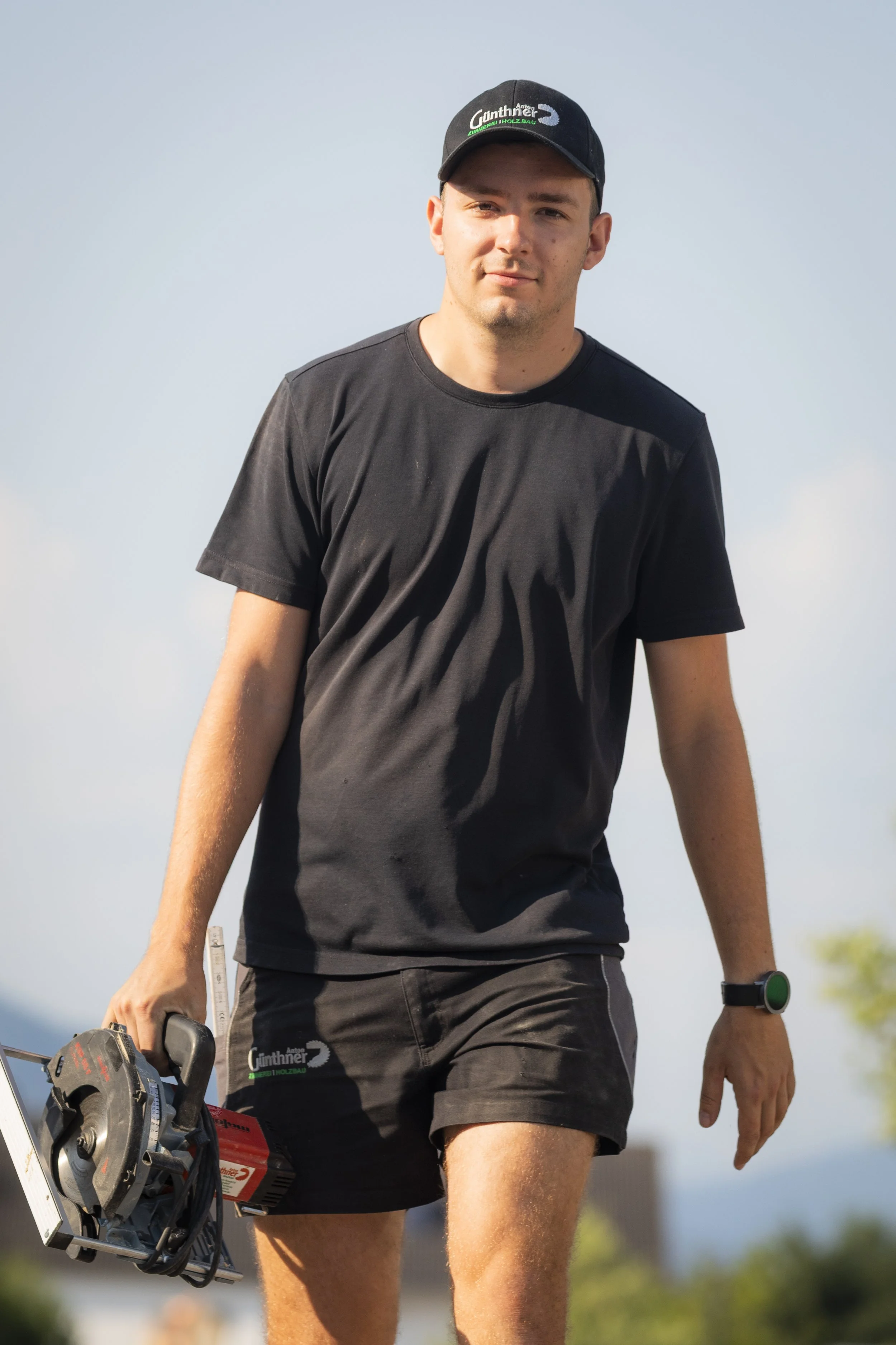 Ein junger Mann in schwarzer Sportkleidung und Mütze hält eine elektrische Säge und einen Wasserwaagearm. Er macht einen Spaziergang im Freien bei sonnigem Wetter.