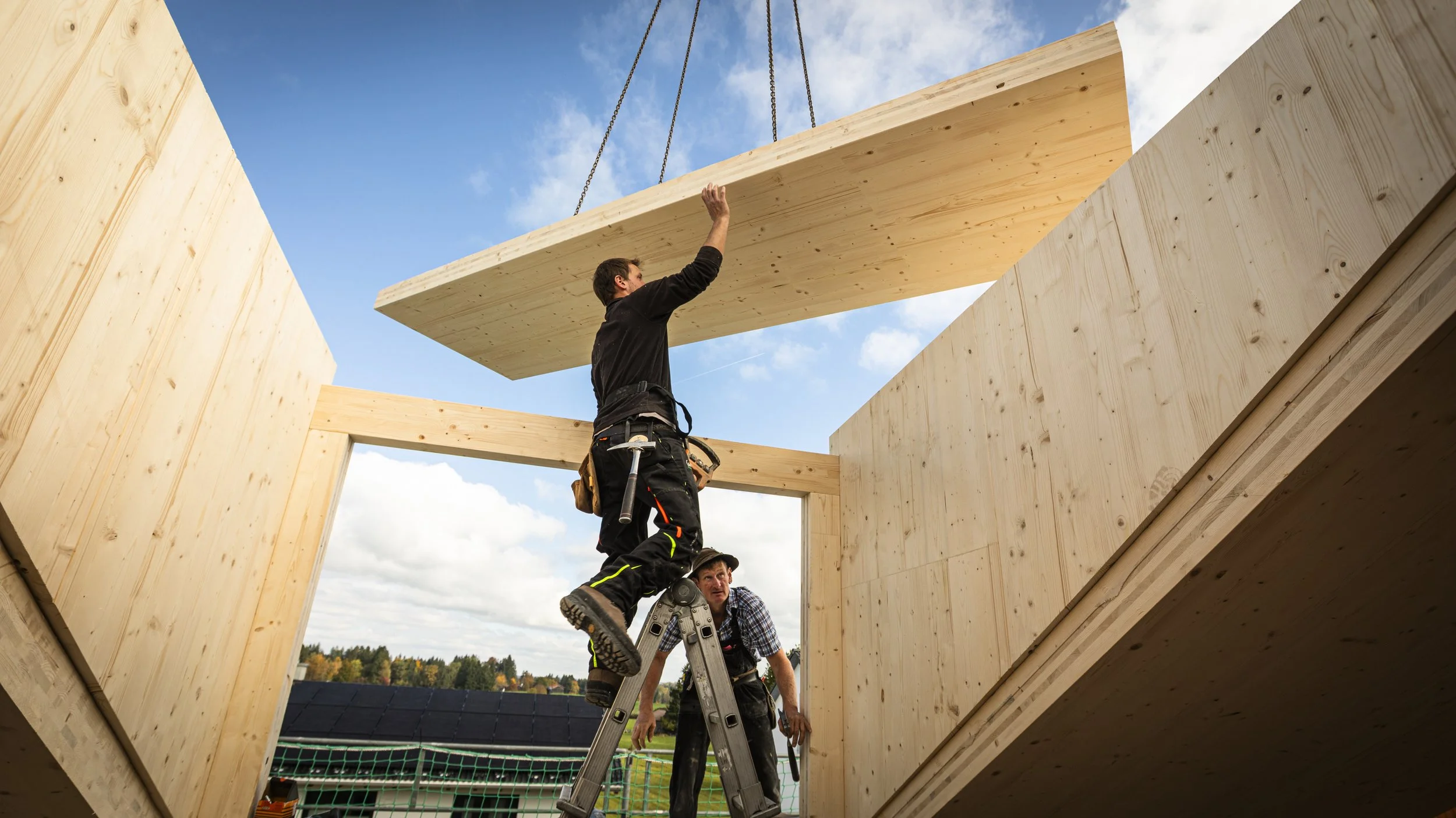 Menschen bauen ein Holzhaus, einer hebt ein großes Holzbrett hoch, während die anderen auf einer Leiter stehen.