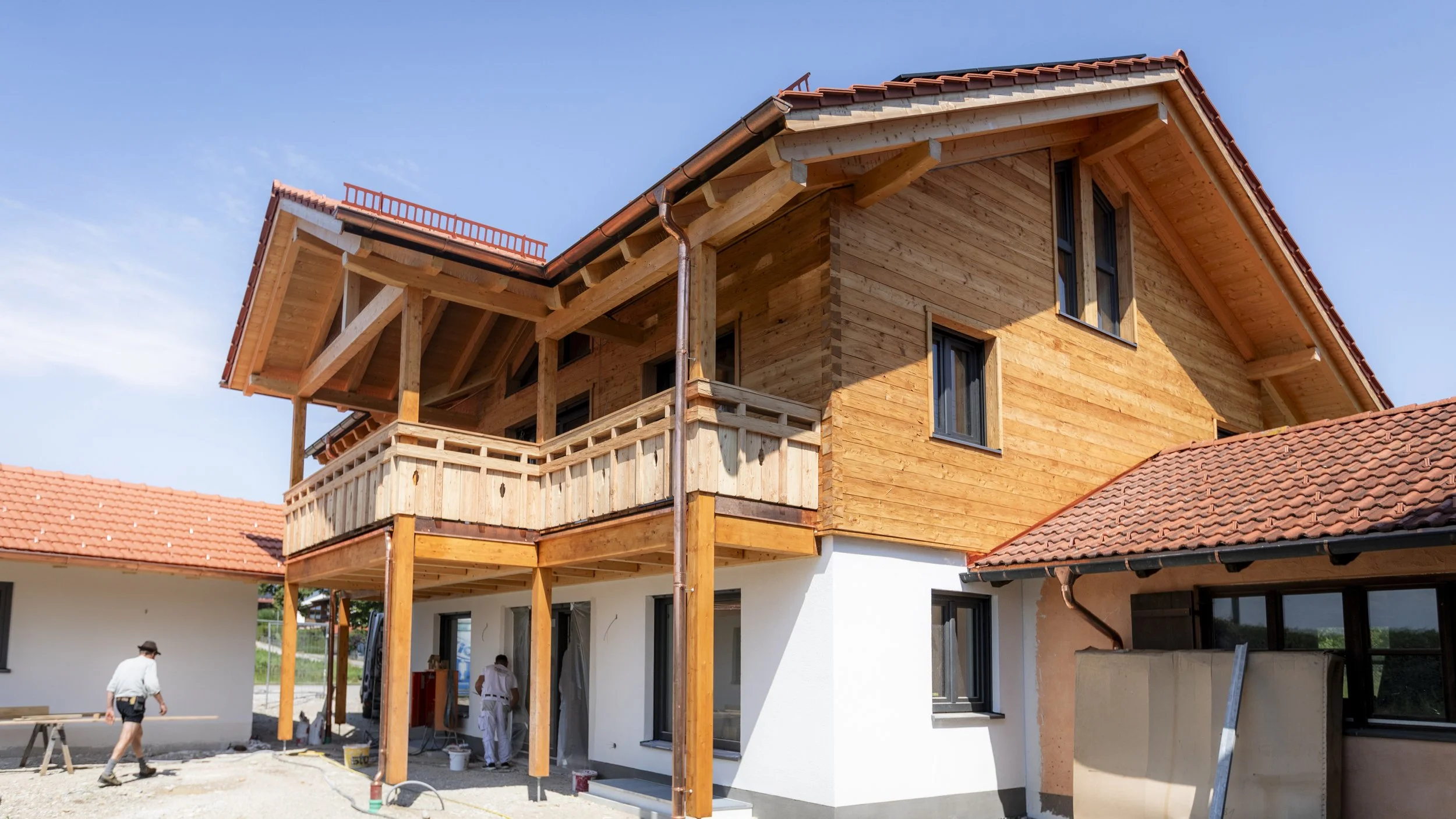 Ein mehrstöckiges Haus mit Holzwänden und roten Ziegeldächern, im Bau befindlich, mit Arbeitern im Vordergrund.