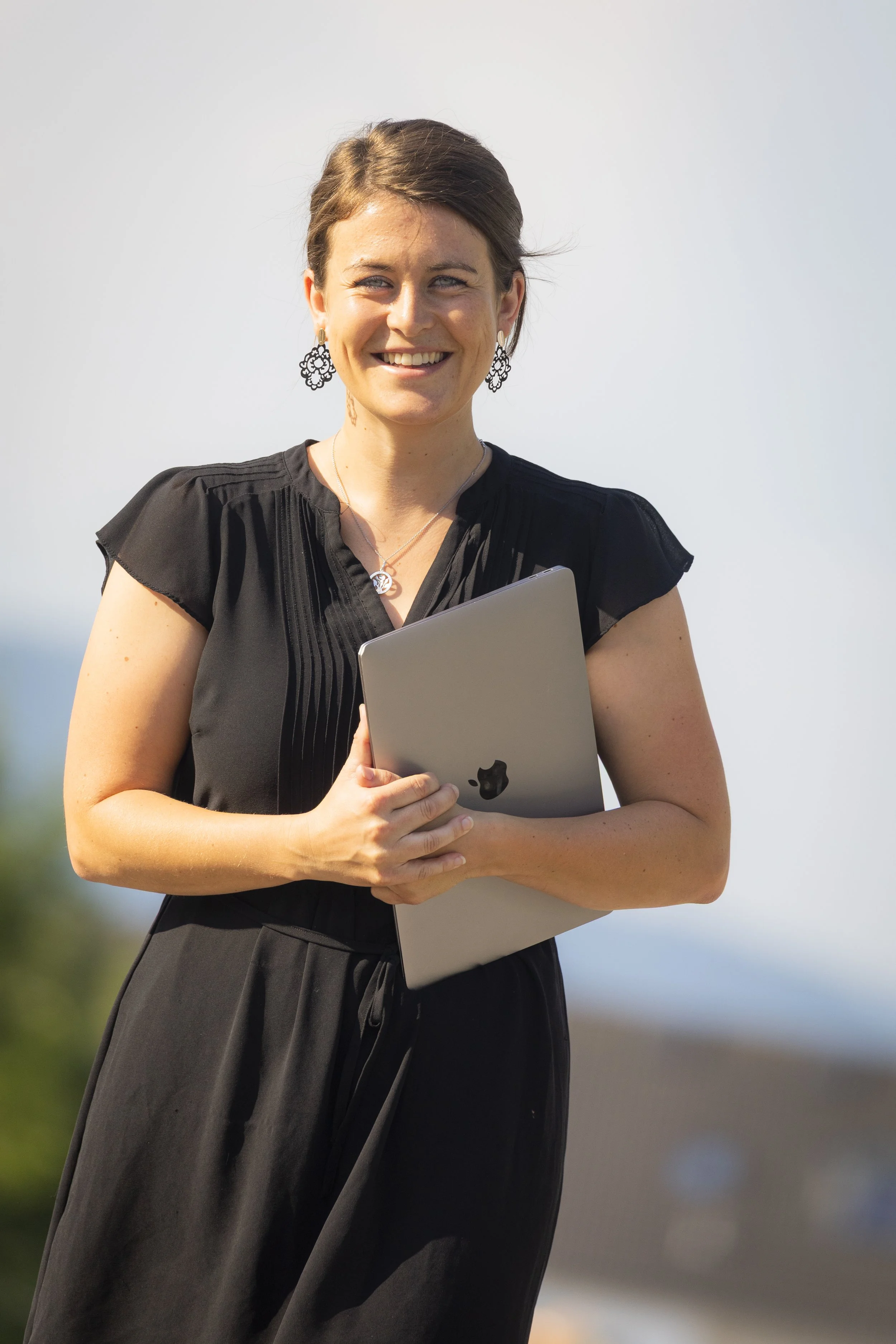Eine lächelnde Frau mit dunklem Haar trägt eine schwarze Kleidung und hält ein MacBook in den Armen, im Hintergrund Himmel und Natur.