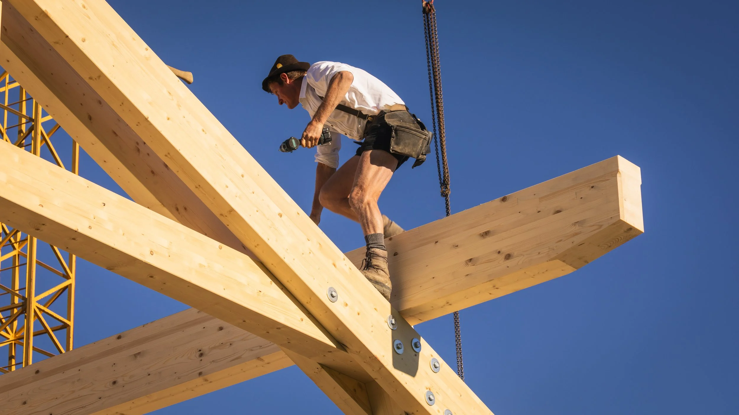 Bauarbeiter auf einer Baustelle, der eine Holzdecke installiert, trägt Schutzkleidung und benutzt ein Werkzeug, während im Hintergrund ein Kran sichtbar ist.
