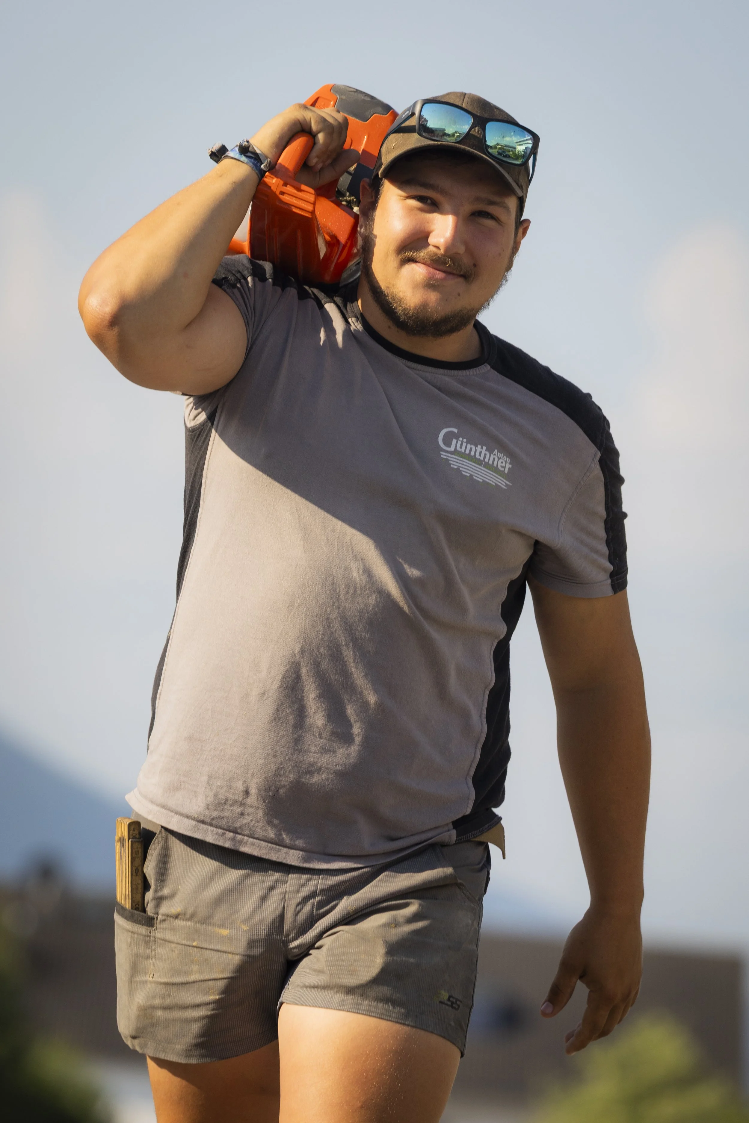 Junge Person trägt eine Bohrmaschine auf der Schulter, trägt eine Cap und Sonnenbrille, trägt ein T-Shirt und Shorts, in einer Outdoor-Umgebung.