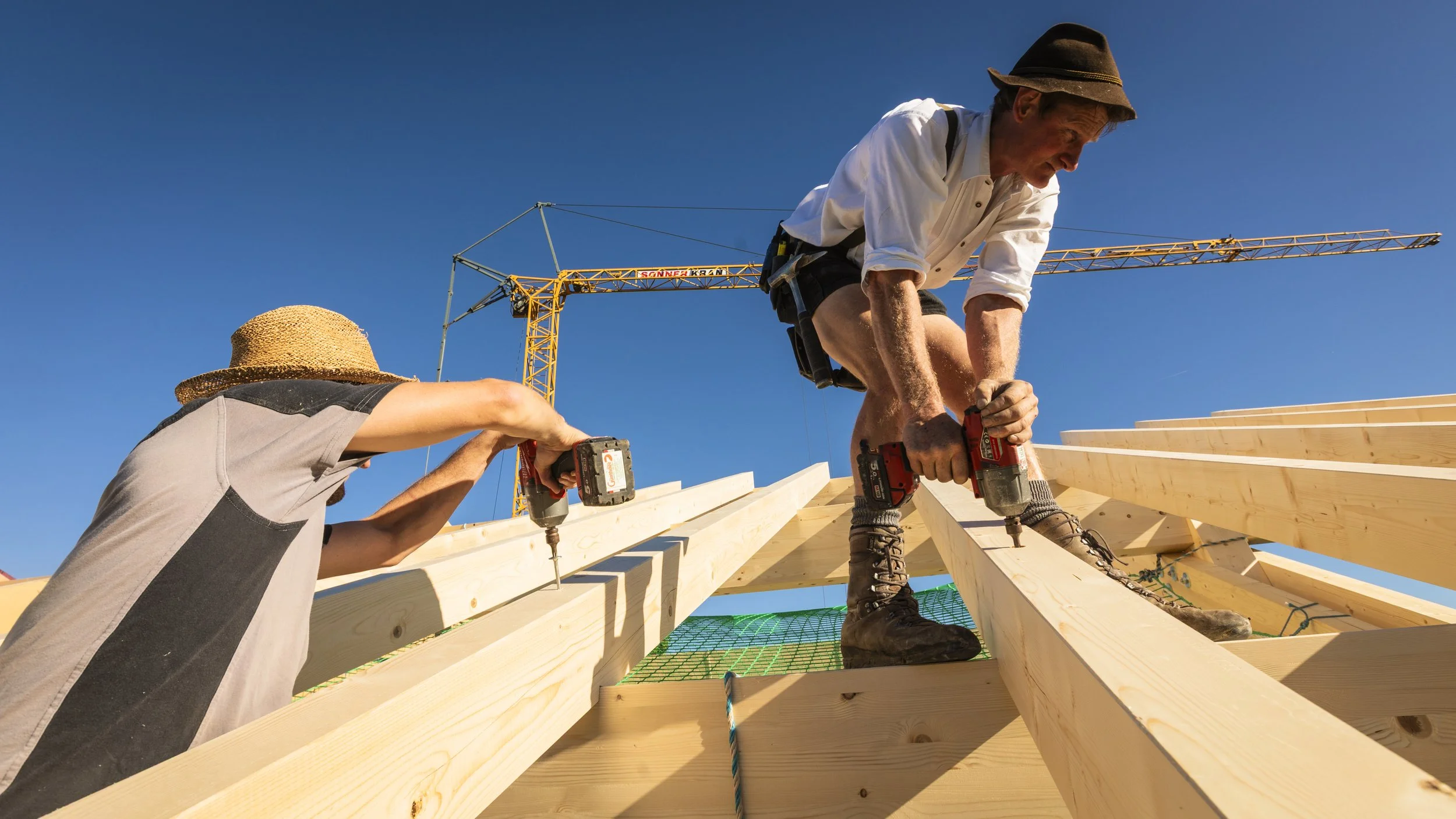 Zwei Männer bauen ein Holzgerüst auf einer Baustelle bei blauem Himmel, einer trägt einen Hut, der andere arbeitet mit einem Akku-Bohrschrauber.