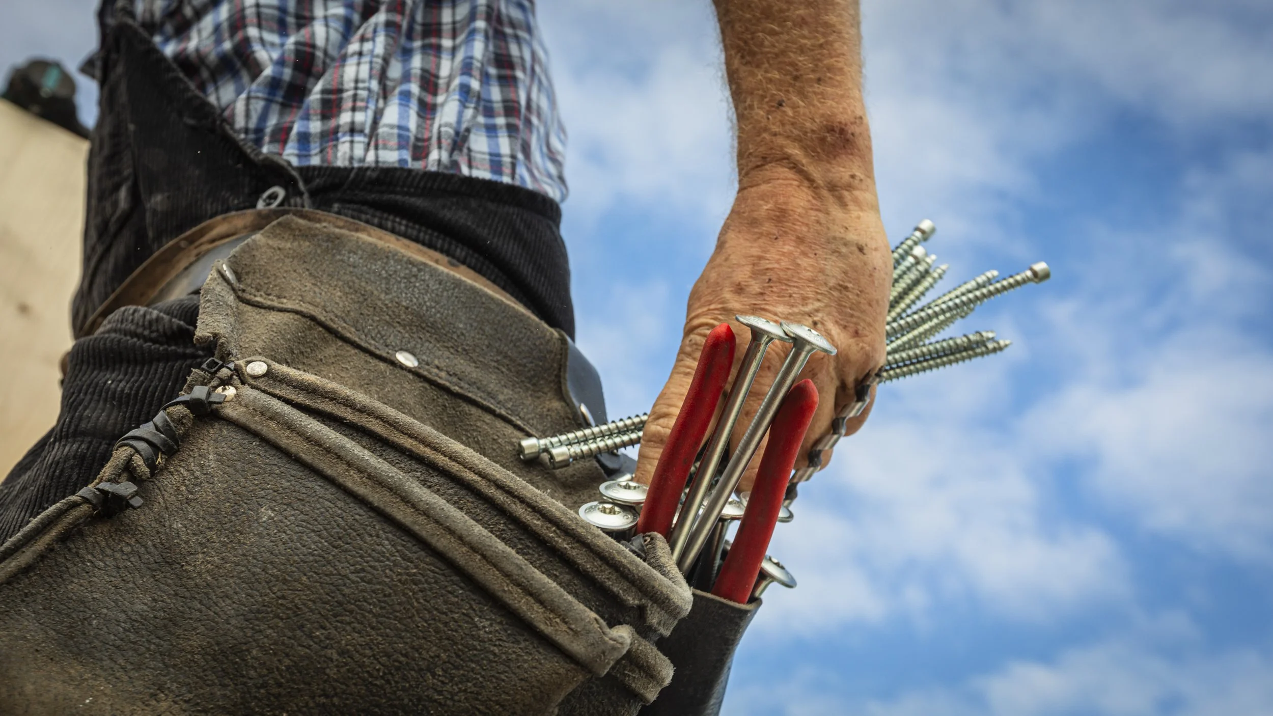 Handwerker mit Werkzeug und Schrauben in der Hand, trägt Arbeitshose und Werkzeugtasche, Himmel im Hintergrund.