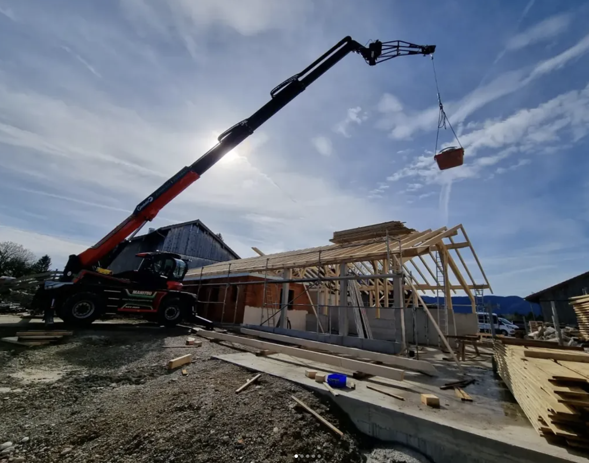 Auf einer Baustelle wird ein Holzrahmen für ein Gebäude errichtet. Ein Kran hebt Baumaterialien, die in einem Korb suspendiert sind.