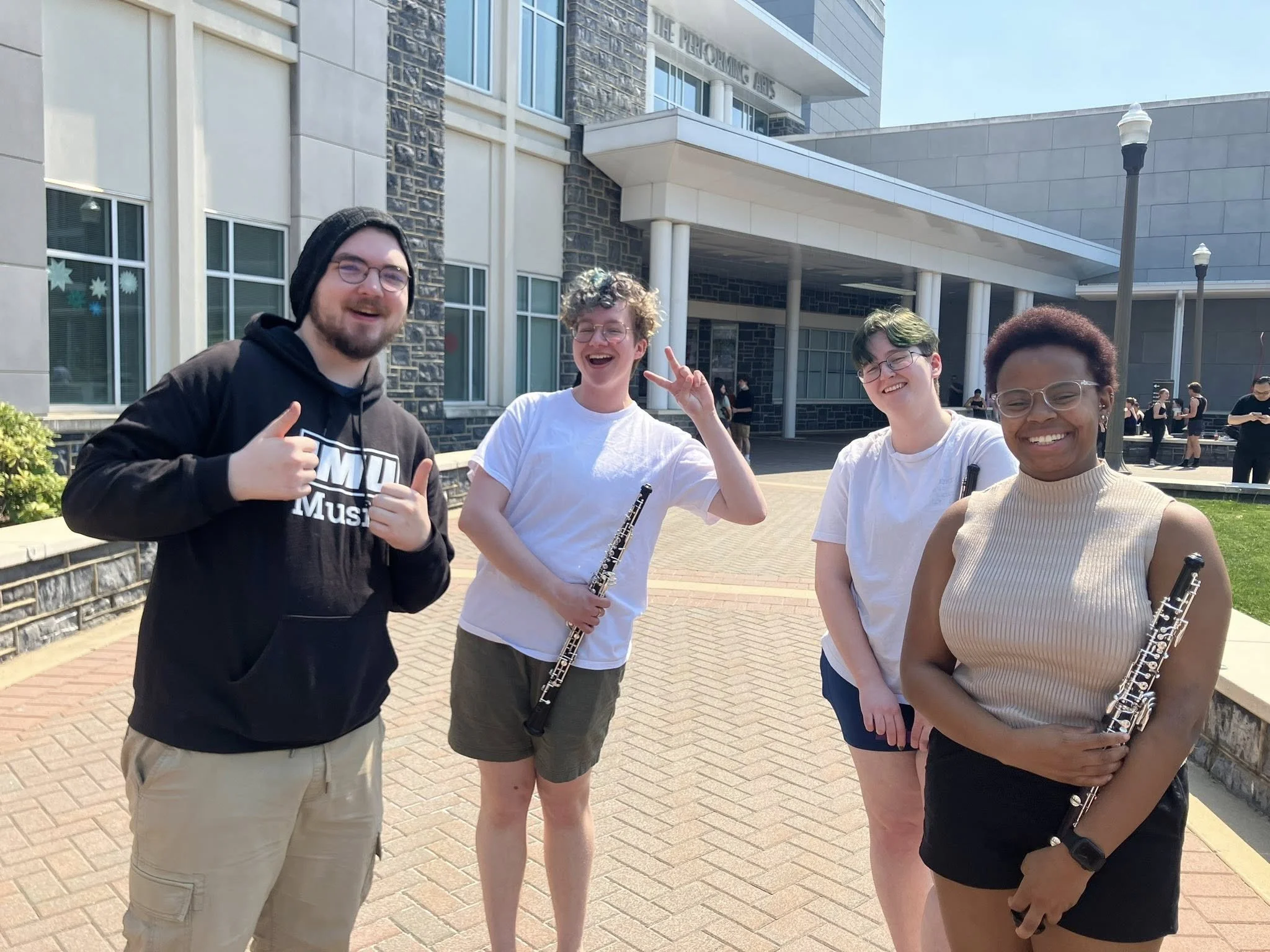 Four people standing outdoors in front of a building, smiling and holding musical instruments.