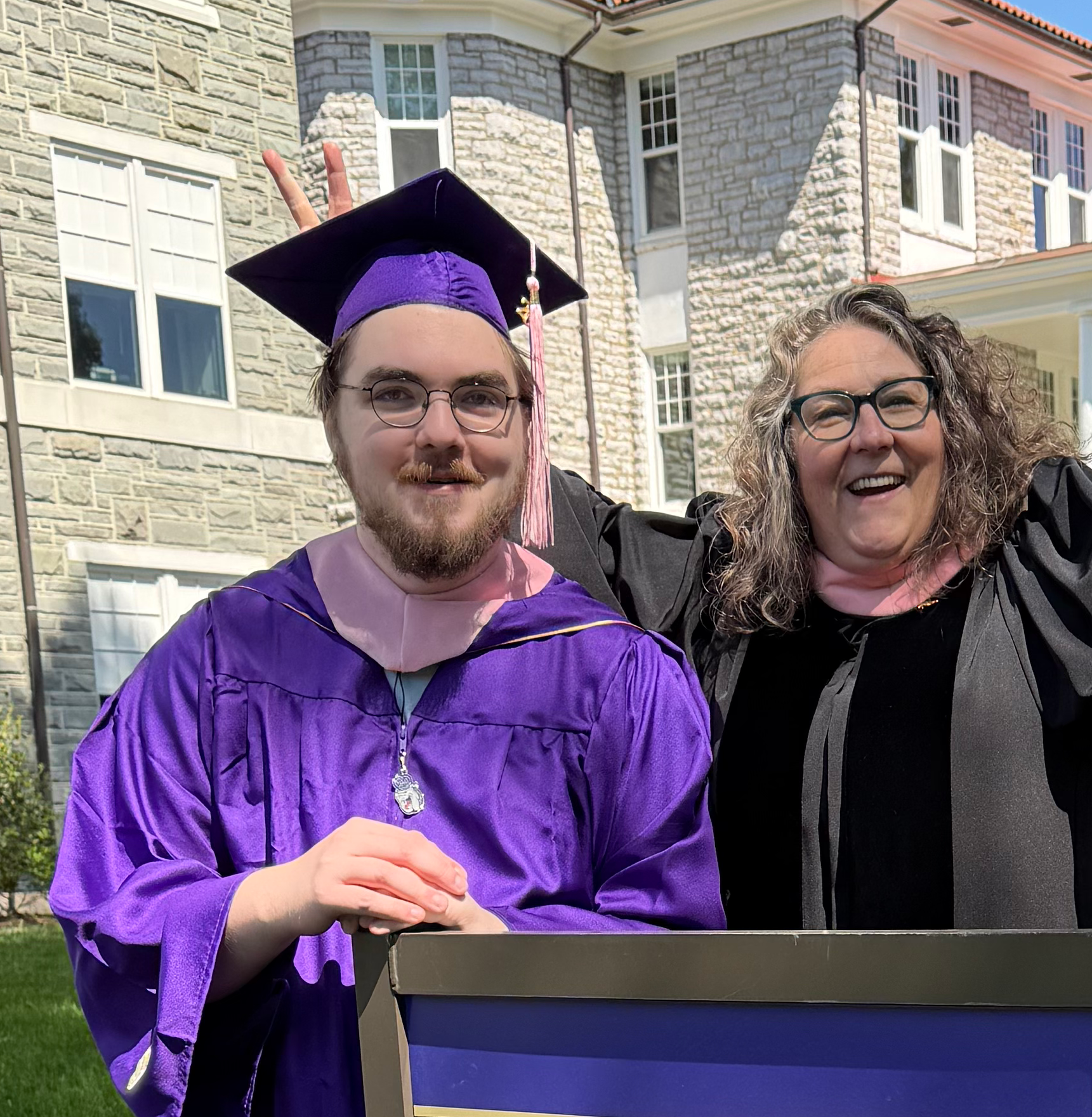 A man in a purple graduation gown and cap making a peace sign behind a woman with curly hair and glasses. They are outdoors in front of a stone house with multiple windows.