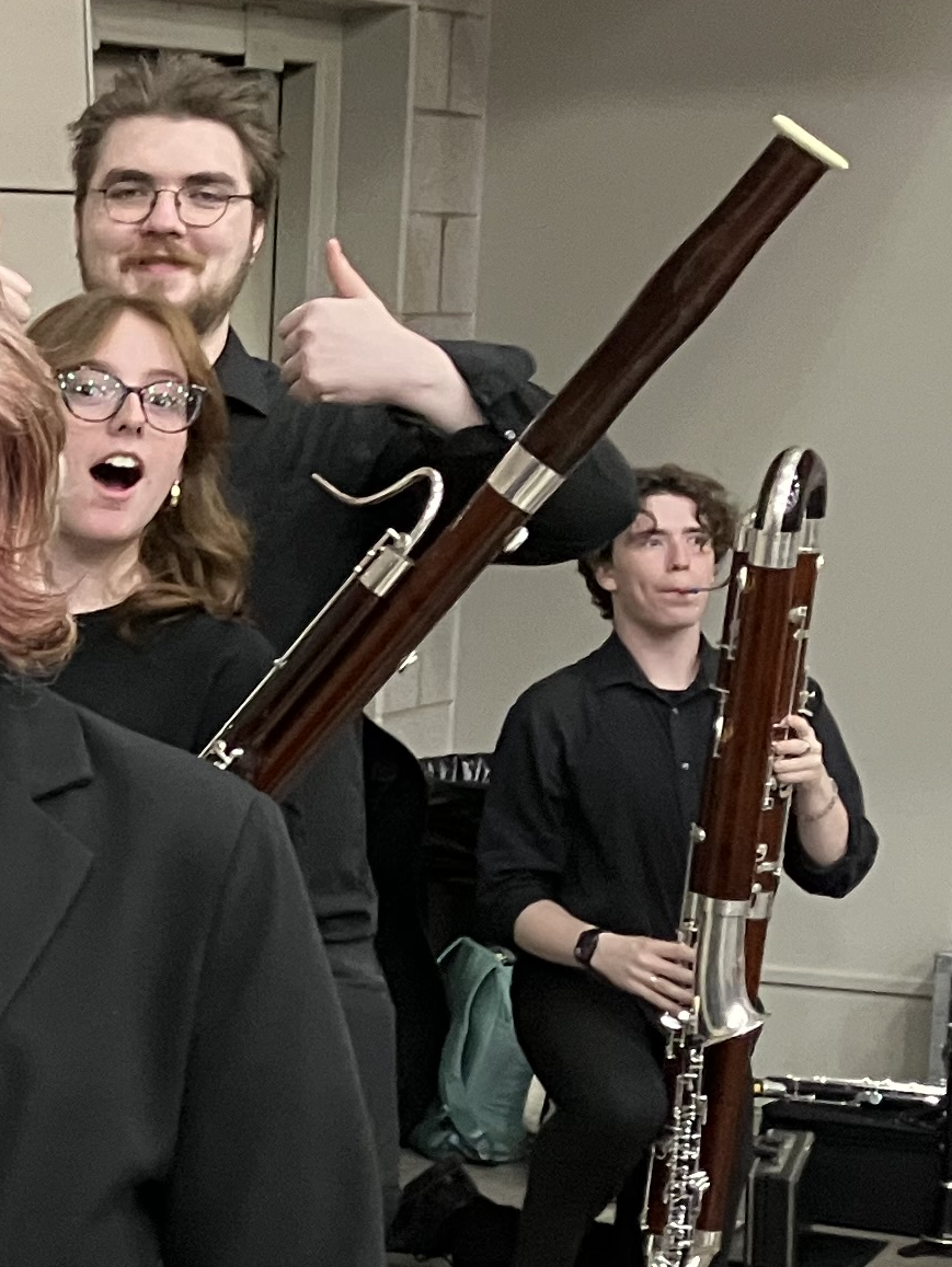 Two people holding bassoons, one giving a thumbs-up and smiling, the other sitting and holding a bassoon, with surprised or amused expressions.