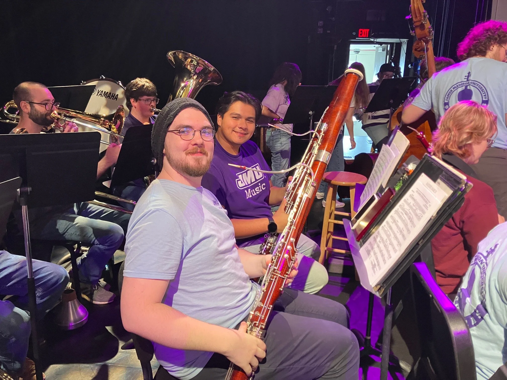 Group of musicians sitting backstage, holding and playing various instruments including a bassoon and trumpets, preparing for a performance.