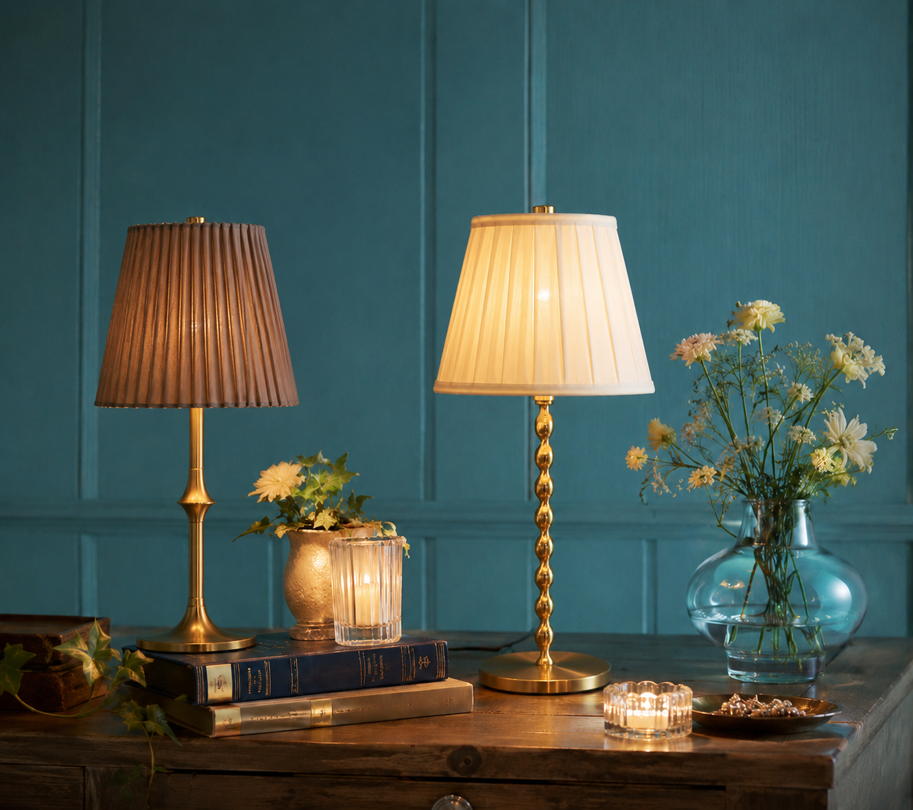 Decorative table with two table lamps, a vase with white flowers, candles, books, and jewelry on a wooden surface against a teal wall.