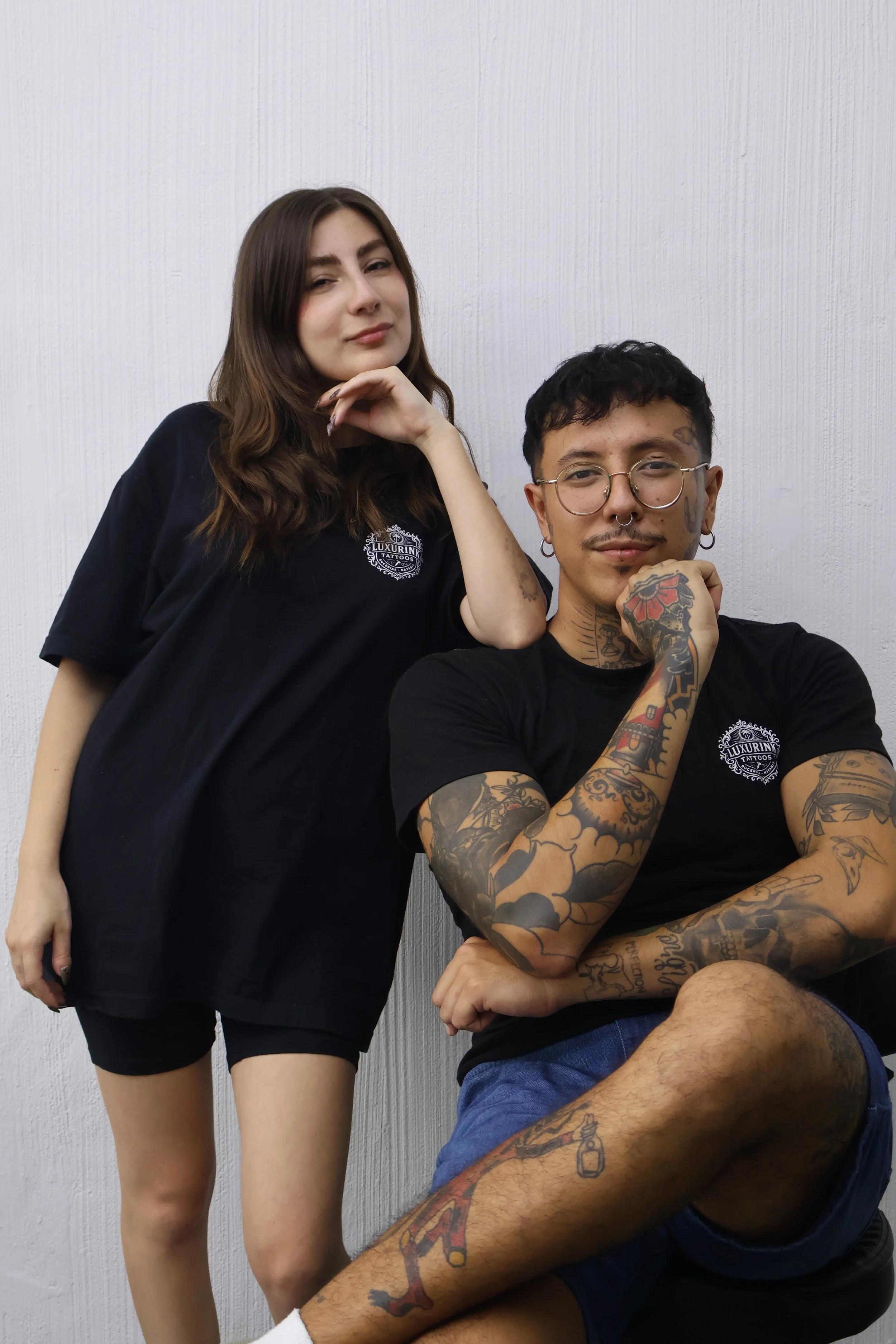 A young woman with brown hair and a young man with tattoos and glasses posing against a white wall. The woman is leaning on the man's shoulder, and the man has his hand near his face.