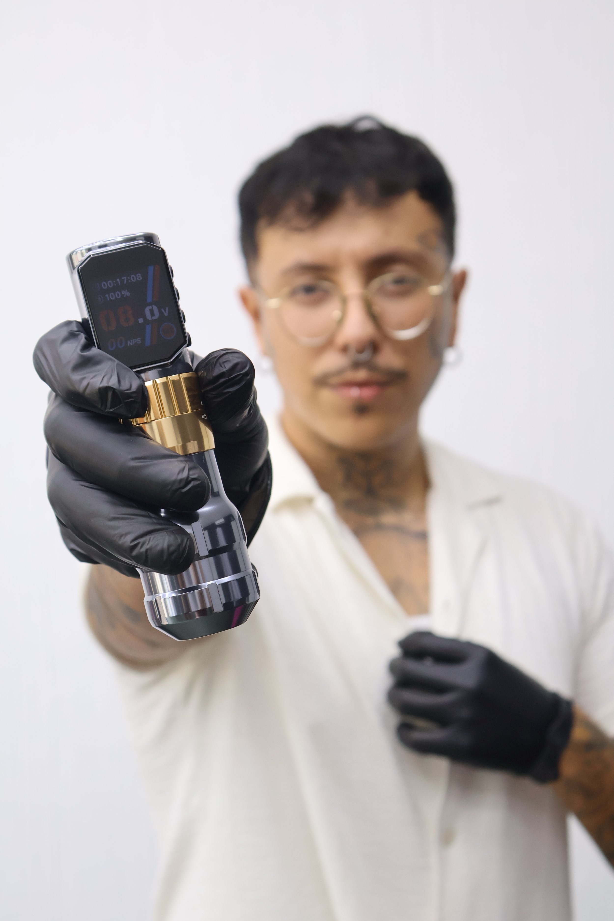 A person with glasses and tattoos holding a black and gold tattoo machine, with a tattoo needle in focus and the person's face blurred in the background.