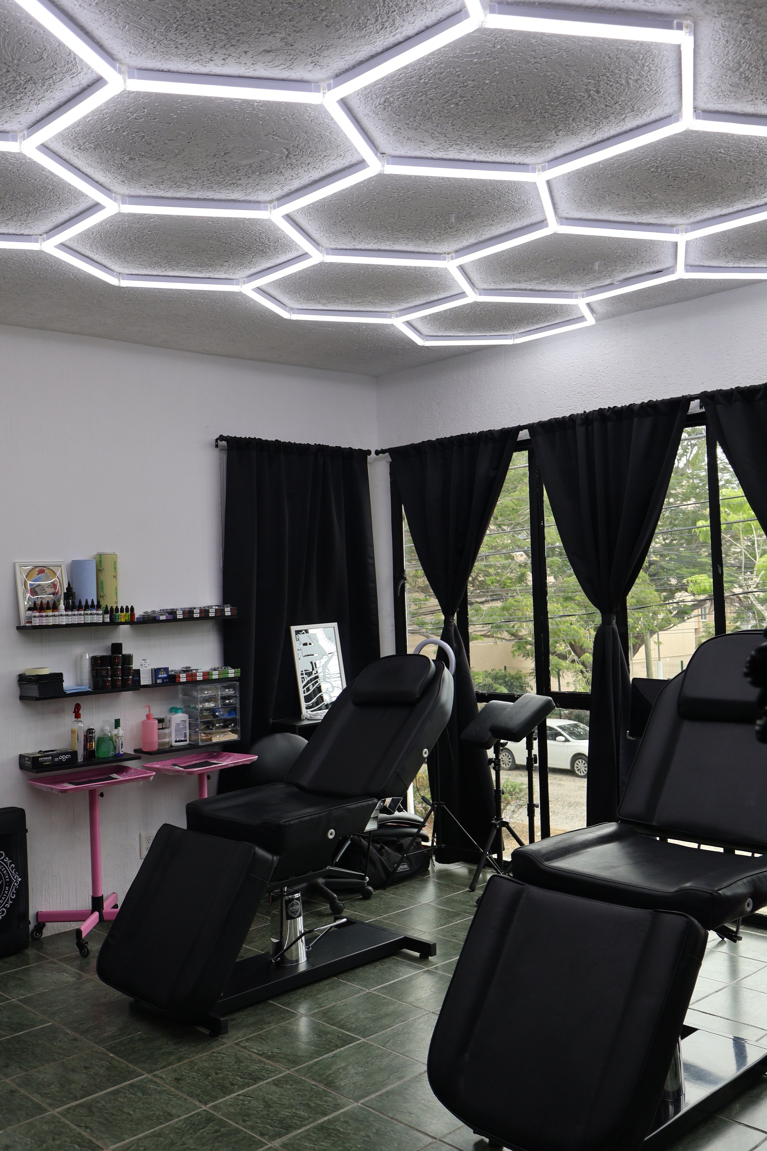 Interior of a tattoo or beauty salon featuring reclining chairs, black curtains, and shelves with nail polish and beauty supplies. Large windows with black curtains allow natural light, and hexagonal LED ceiling lights illuminate the space.