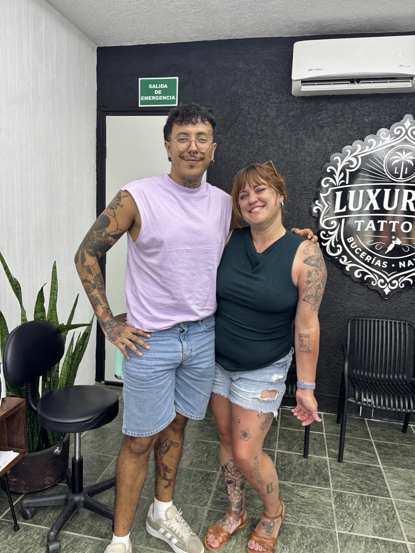 A man and a woman standing inside a tattoo studio, smiling at the camera. The man has glasses, a nose ring, and tattoos on his arms and legs, wearing a light purple sleeveless shirt and denim shorts. The woman has tattoos on her arms and legs, and is wearing a dark sleeveless top and ripped denim shorts. The tattoo studio sign behind them reads 'LUXUR TATTOO,' and there is a green emergency exit sign above a door in the background.