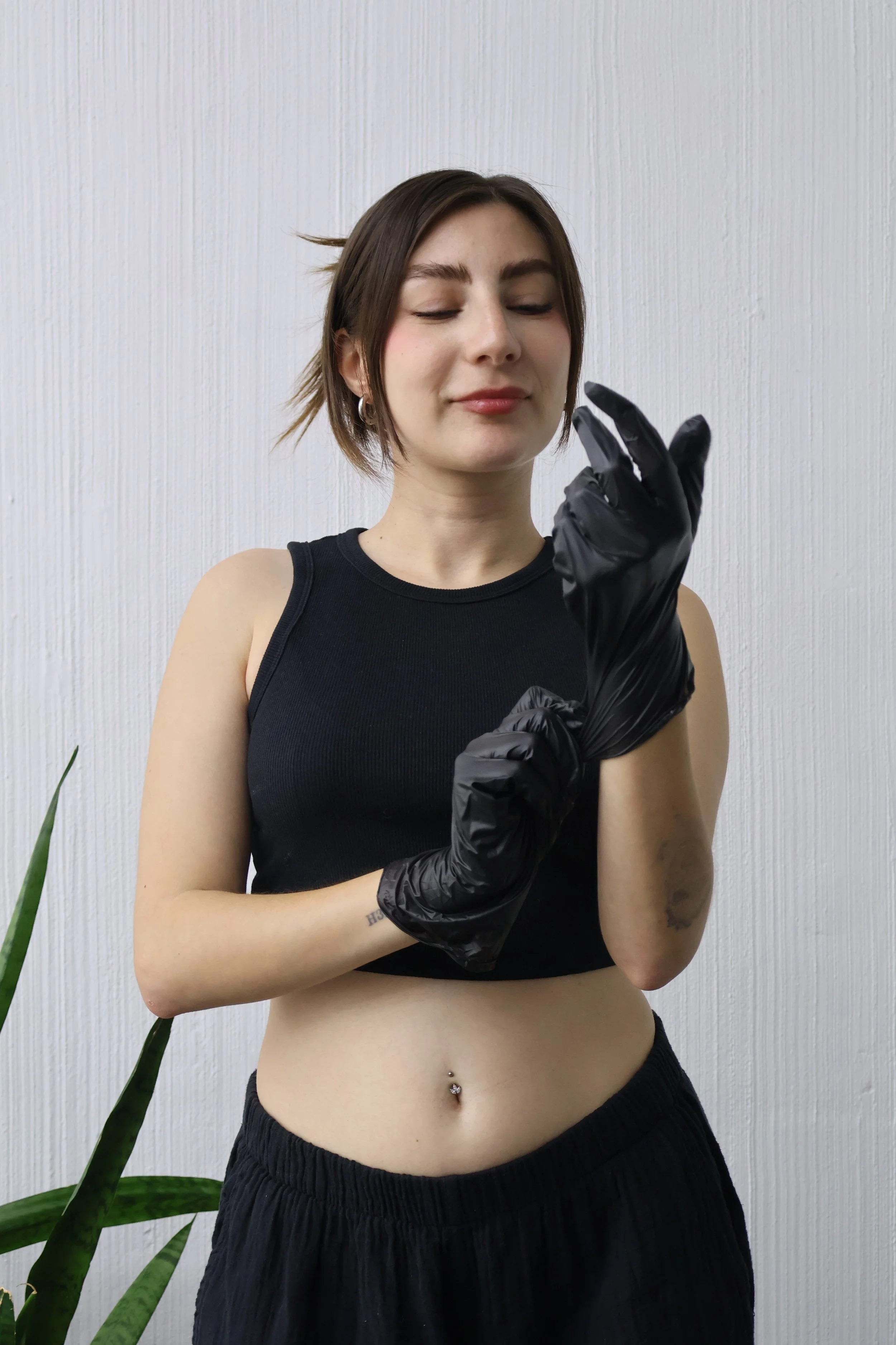 A young woman with short brown hair and fair skin, dressed in a black sleeveless top, wearing black gloves, with a silver belly button piercing, standing against a plain white wall, with a green plant partially visible on the left side.