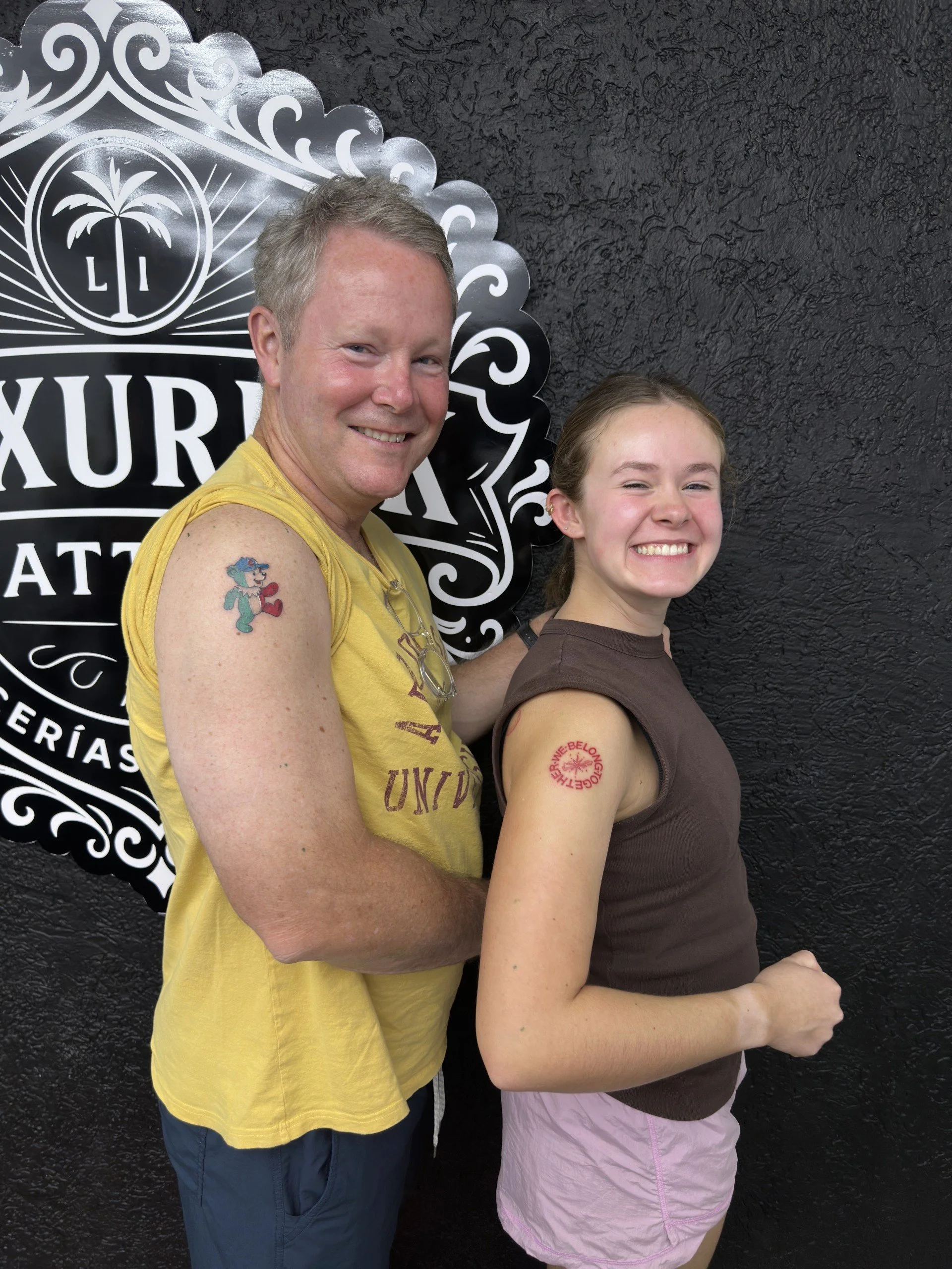 Two smiling people, a man with short gray hair wearing a yellow tank top and a woman with long brown hair wearing a brown shirt and pink shorts, standing side by side in front of a black textured wall with a decorative sign featuring a palm tree and the letters L and I.