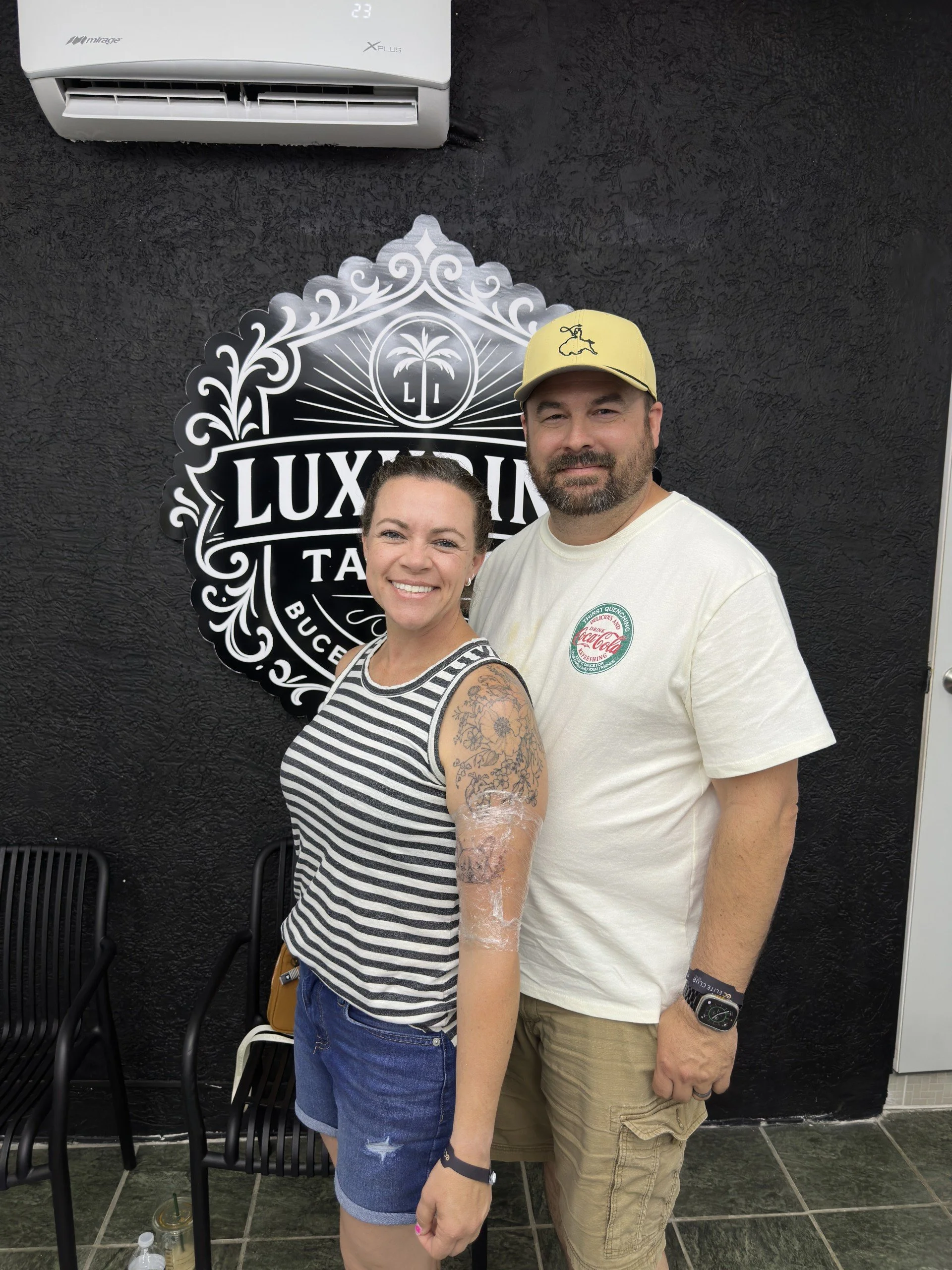 A woman with tattoos on her arm and a striped shirt smiling next to a man with a beard and a yellow cap, standing in front of a black textured wall with a sign that says 'LUXYARD TA' and a white air conditioning unit above.