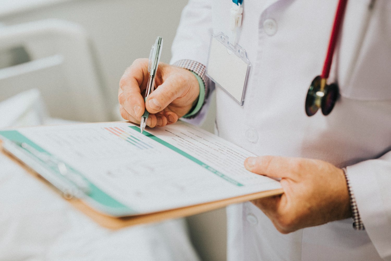 A healthcare professional in a white coat with a stethoscope around their neck is writing on a medical chart or form with a pen.