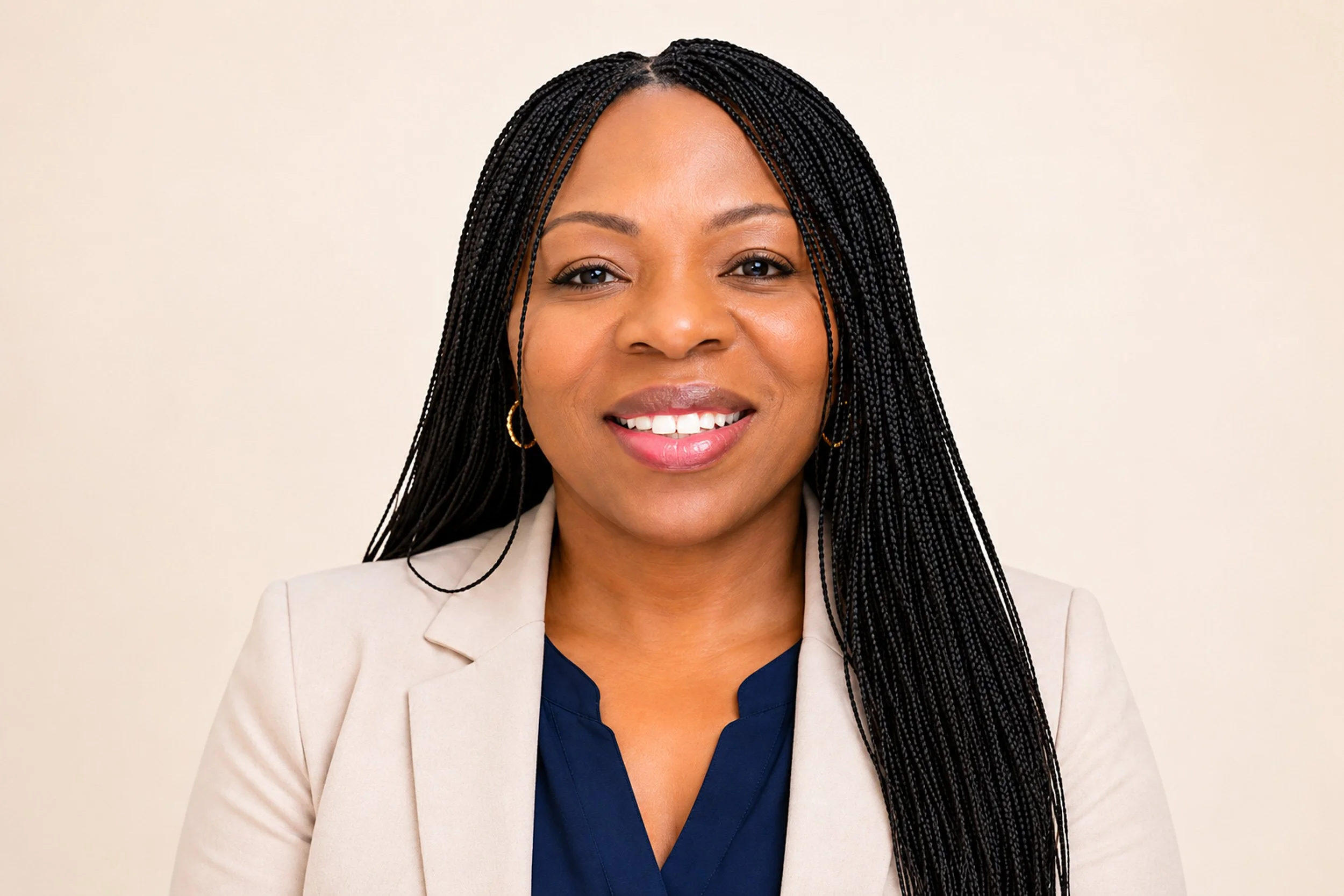 A smiling woman with long black braids, wearing a beige blazer and earrings, against a plain light background.