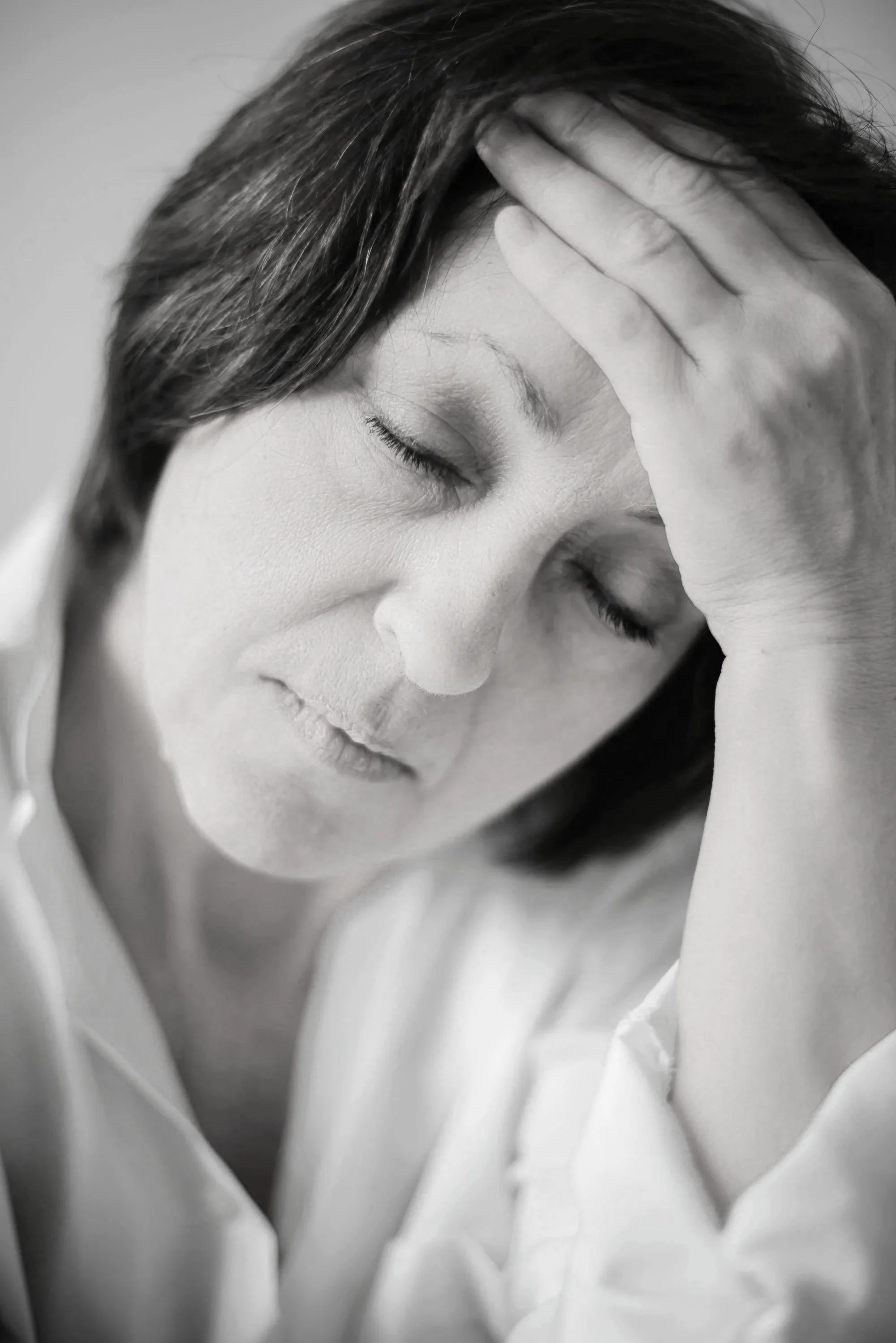 A black and white close-up photo of a woman with short hair holding her forehead with her right hand and eyes closed, appearing distressed.