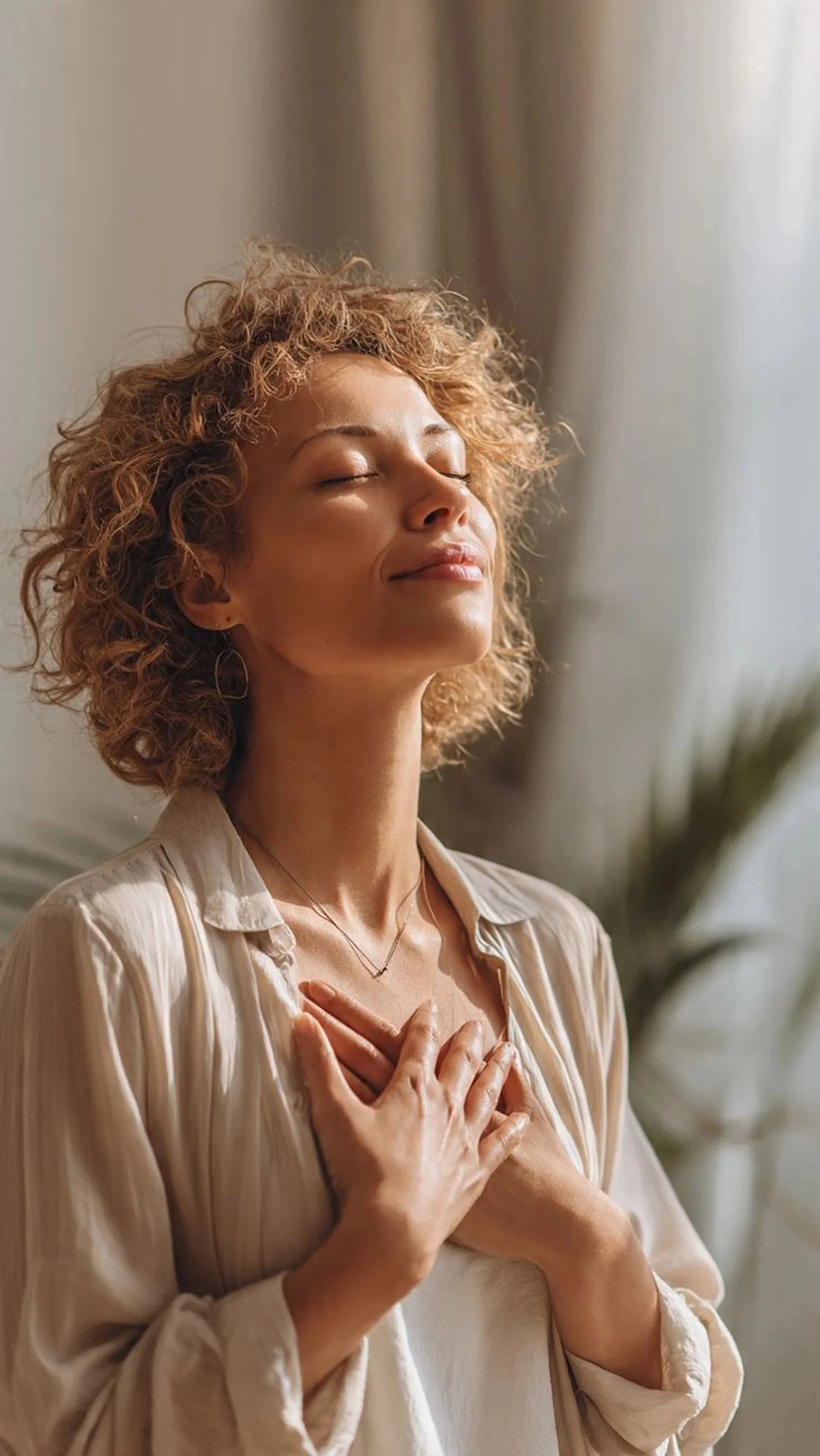 A woman with curly hair, eyes closed, smiling softly, with one hand on her chest and the other on her stomach, standing indoors in front of a neutral background.
