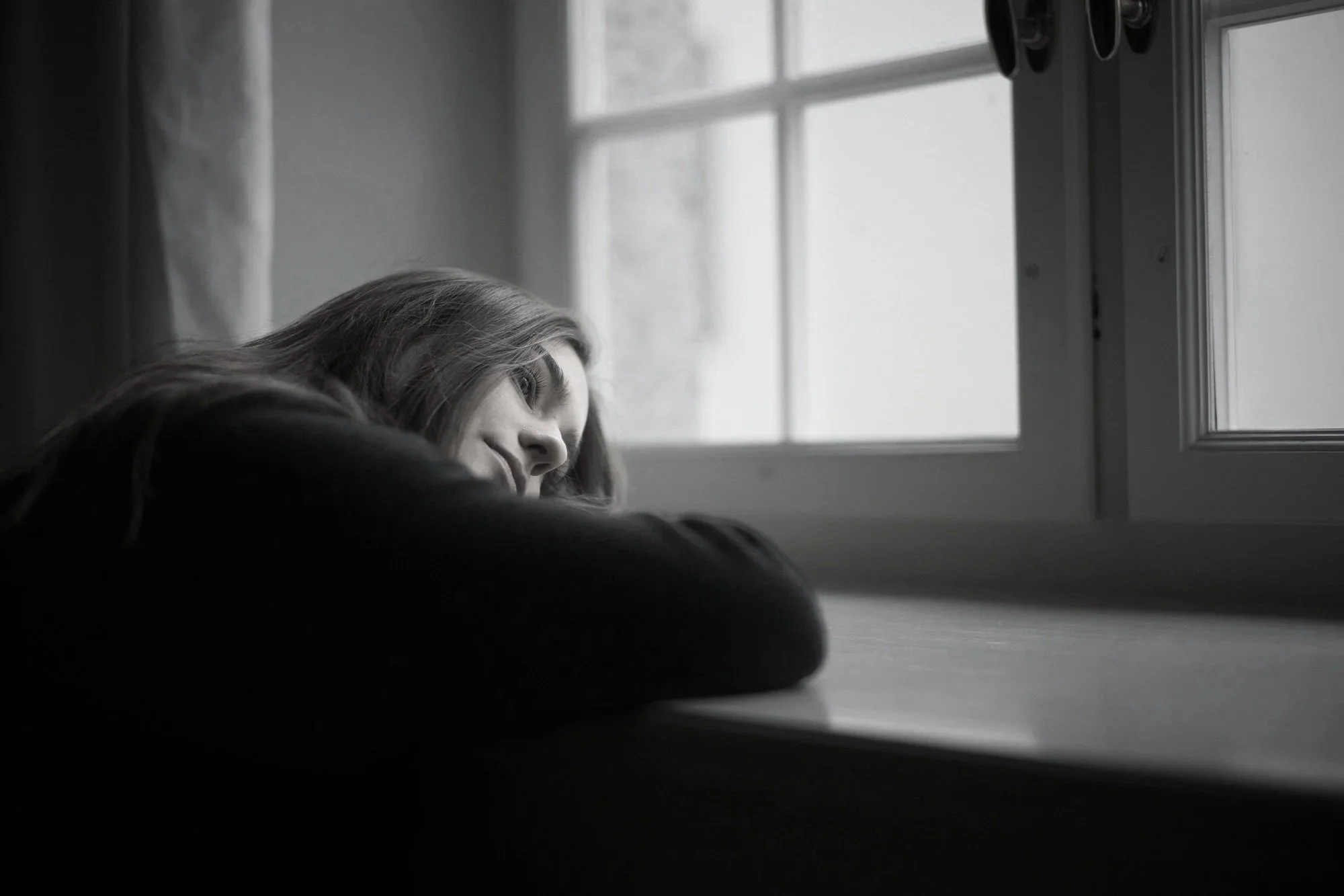 Black and white photo of a woman with long hair resting her head on her arms near a window.