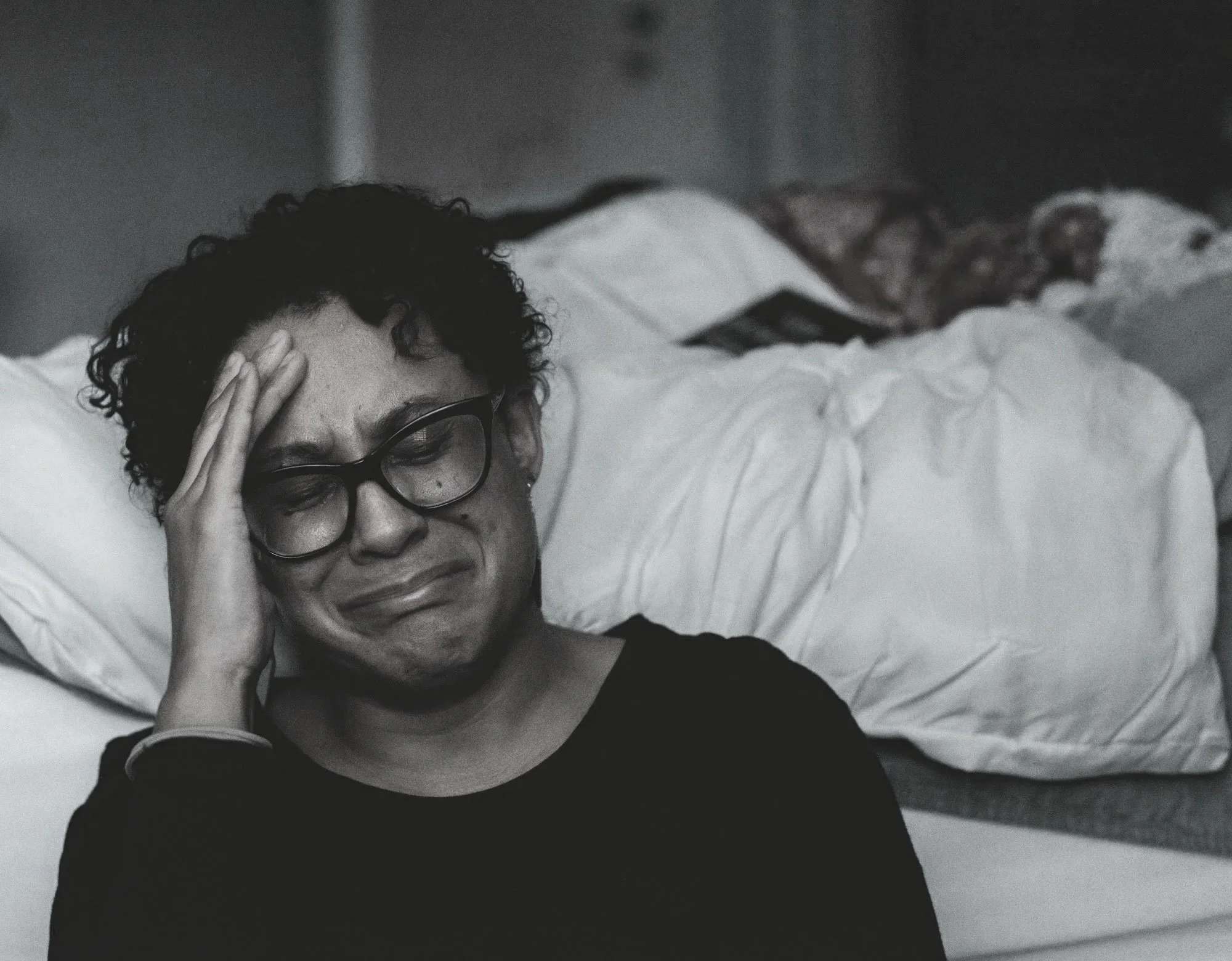 A woman with curly hair and glasses lying in bed, holding her forehead with a distressed expression.