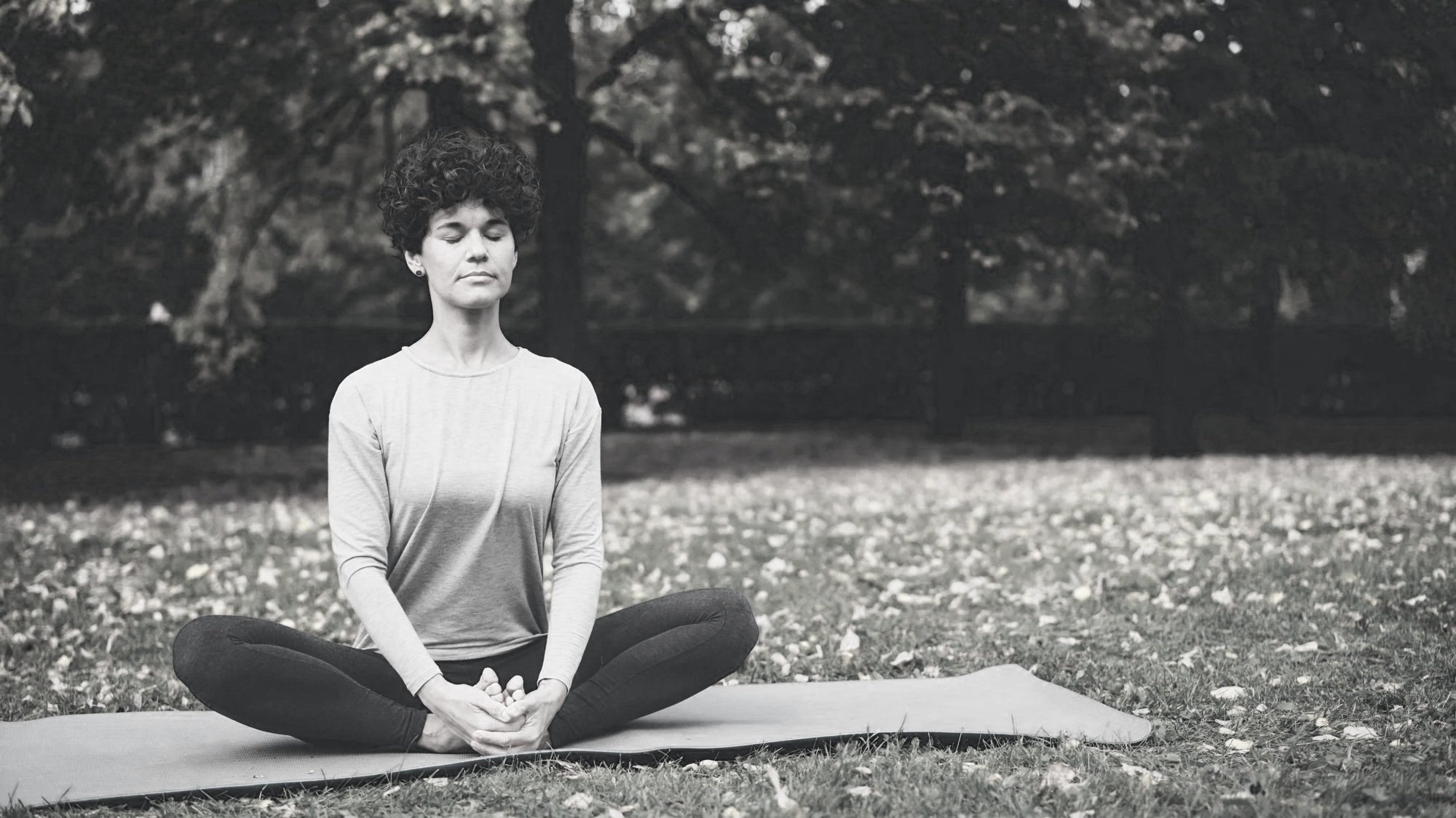 A woman practicing yoga outdoors, sitting cross-legged on a mat with her hands resting on her knees, eyes closed, in a peaceful meditation pose surrounded by fallen leaves in a park with trees in the background.