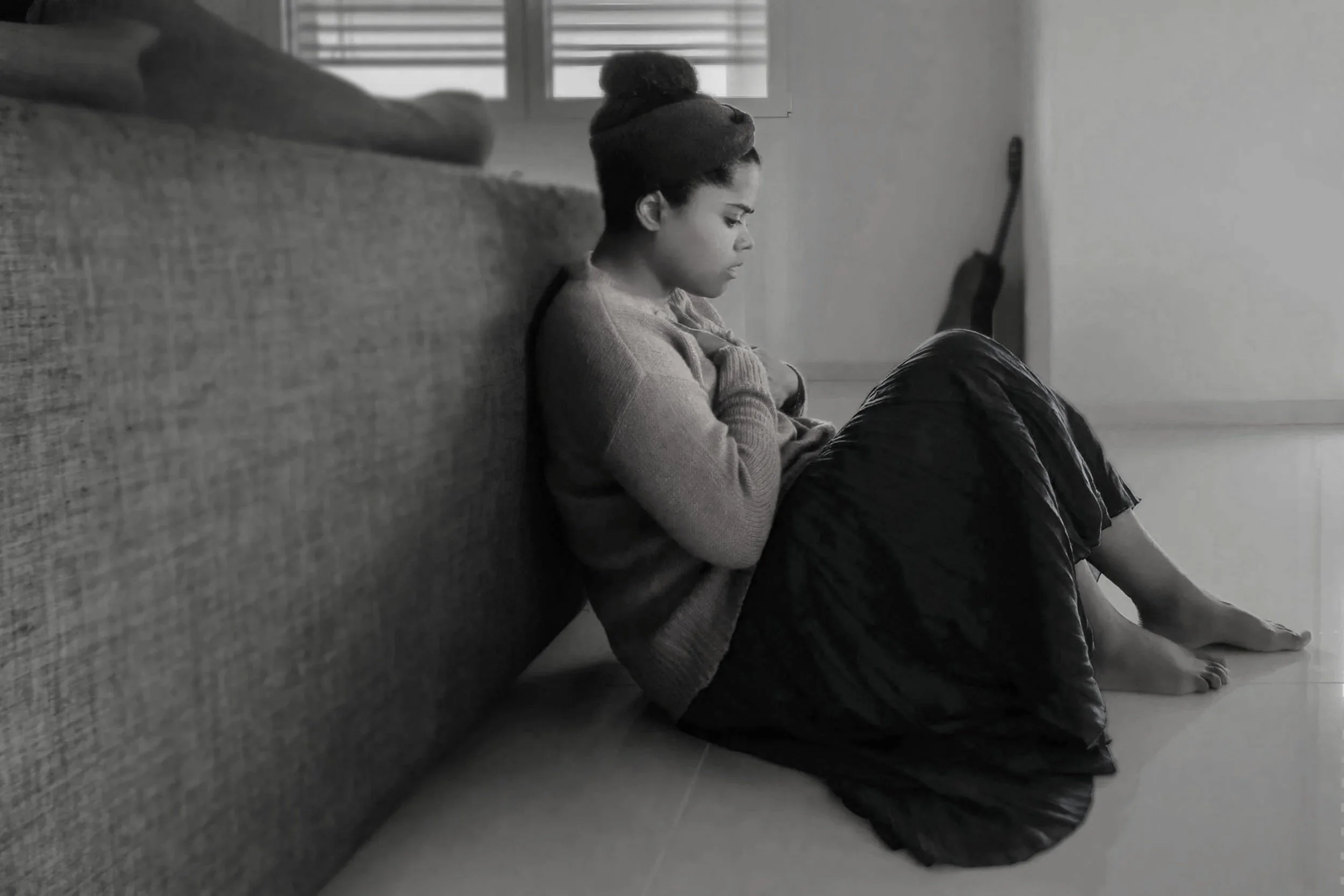 A young girl sitting on the floor next to a couch, looking down with her knees drawn up, in a room with a guitar leaning against the wall.