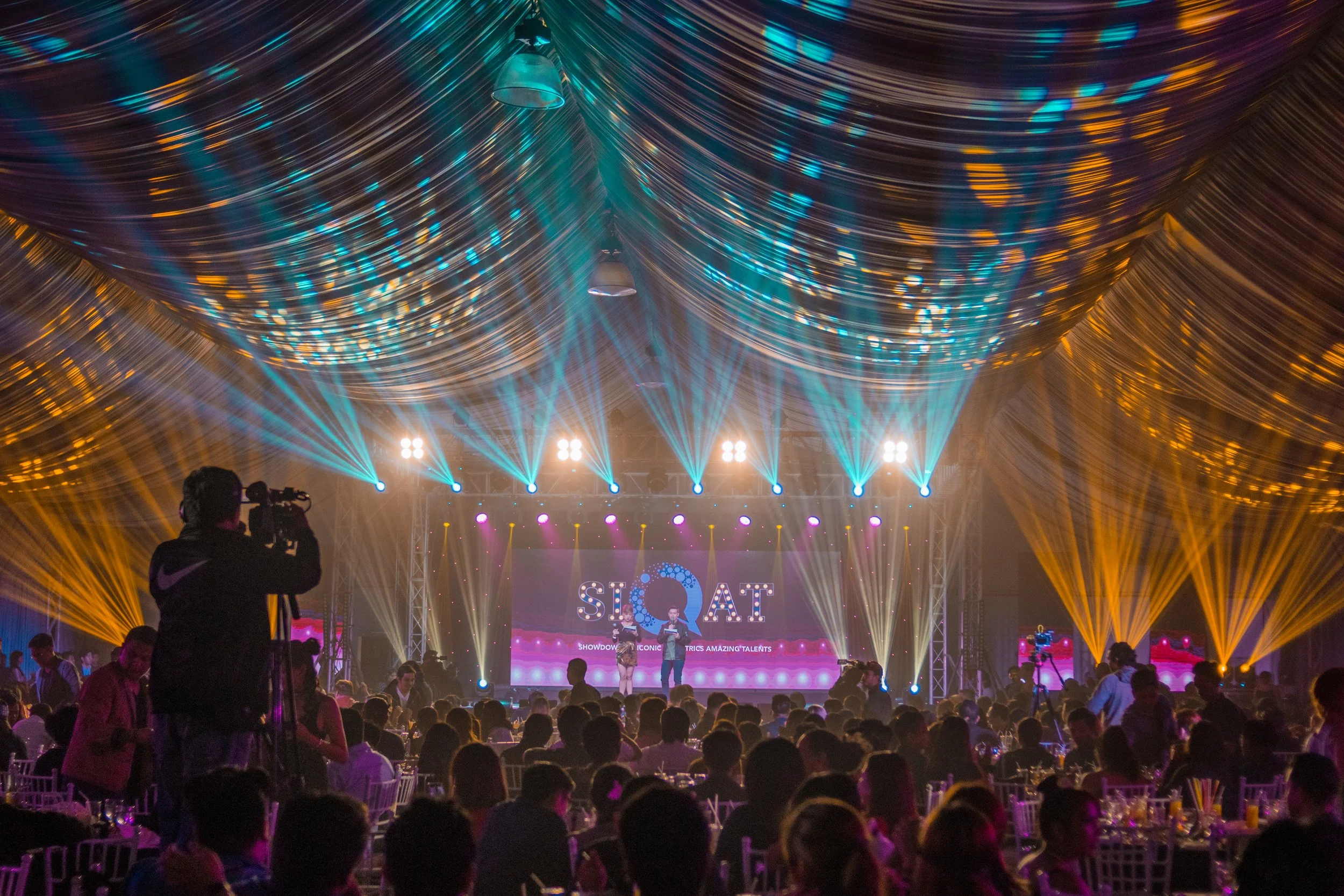 An indoor stage with bright colorful lights and a crowd watching a performance or event, with a large illuminated sign that reads 'SIT...AT' in the background.