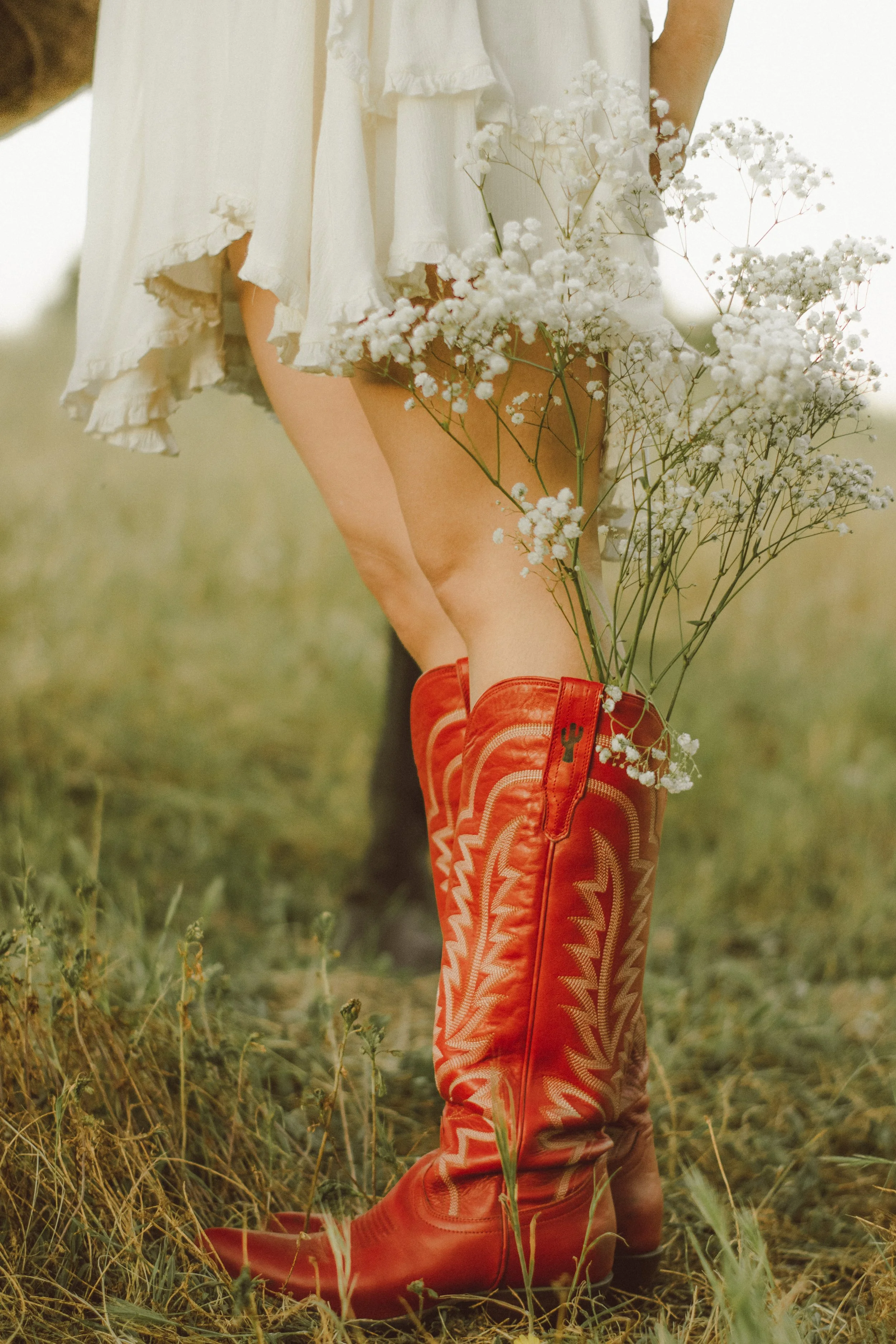 Person wearing red cowboy boots and a white dress holding white baby's breath flowers in a grassy field.