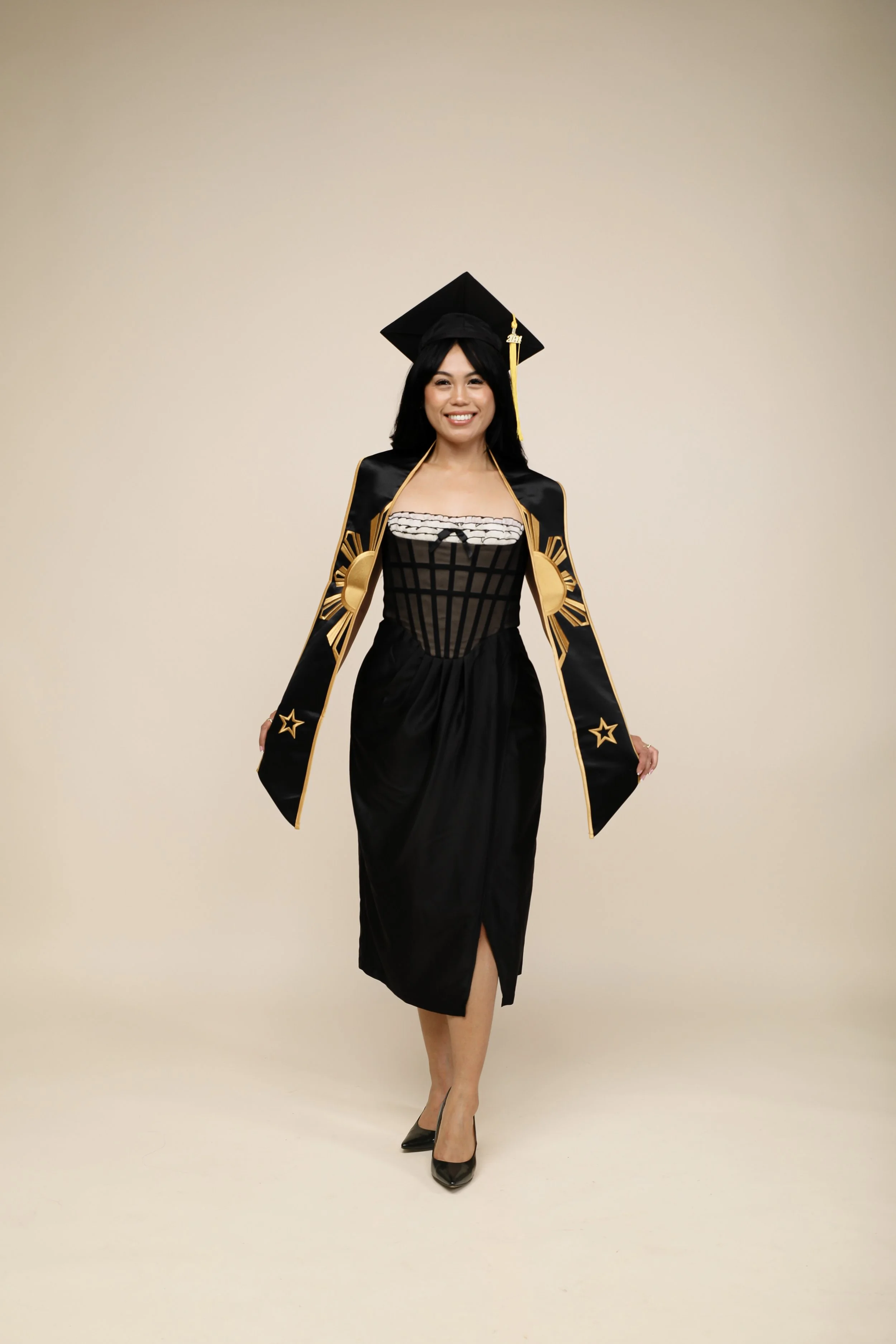 A woman in a black dress and high heels holding a graduation gown and cap, standing against a plain beige background.
